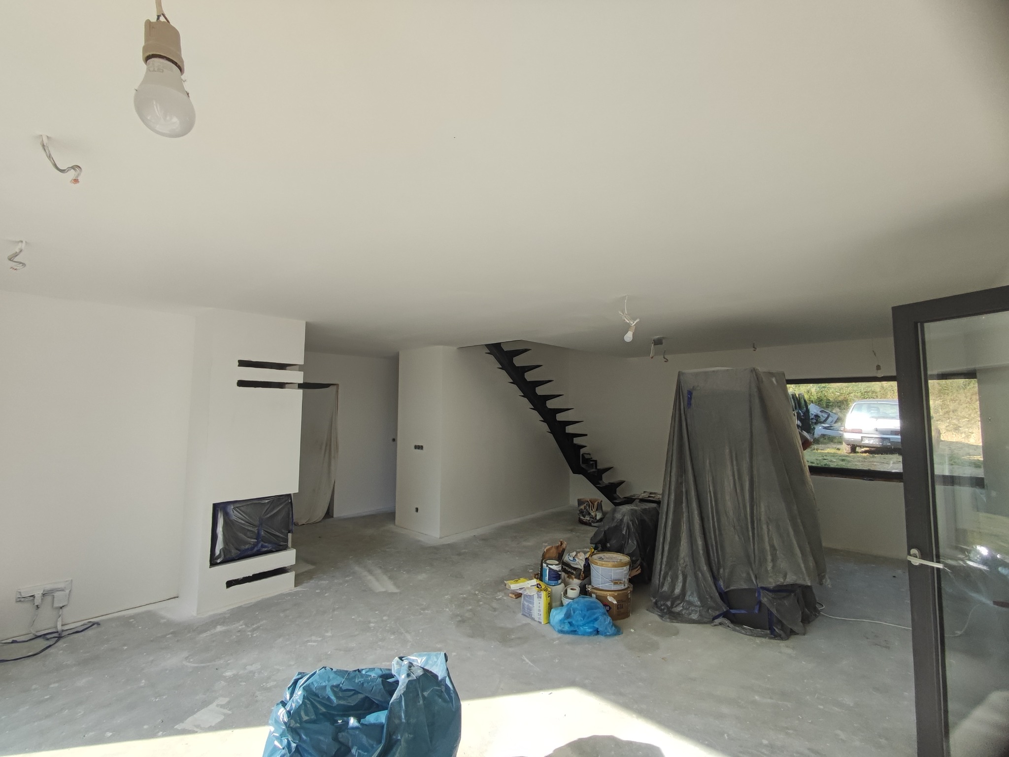 Wnętrze w trakcie prac wykończeniowych: białe ściany, surowa podłoga, zakryty folią mebel, widoczne puszki z farbą i narzędzia, nowoczesny kominek z czarnym wkładem zabezpieczonym folią.