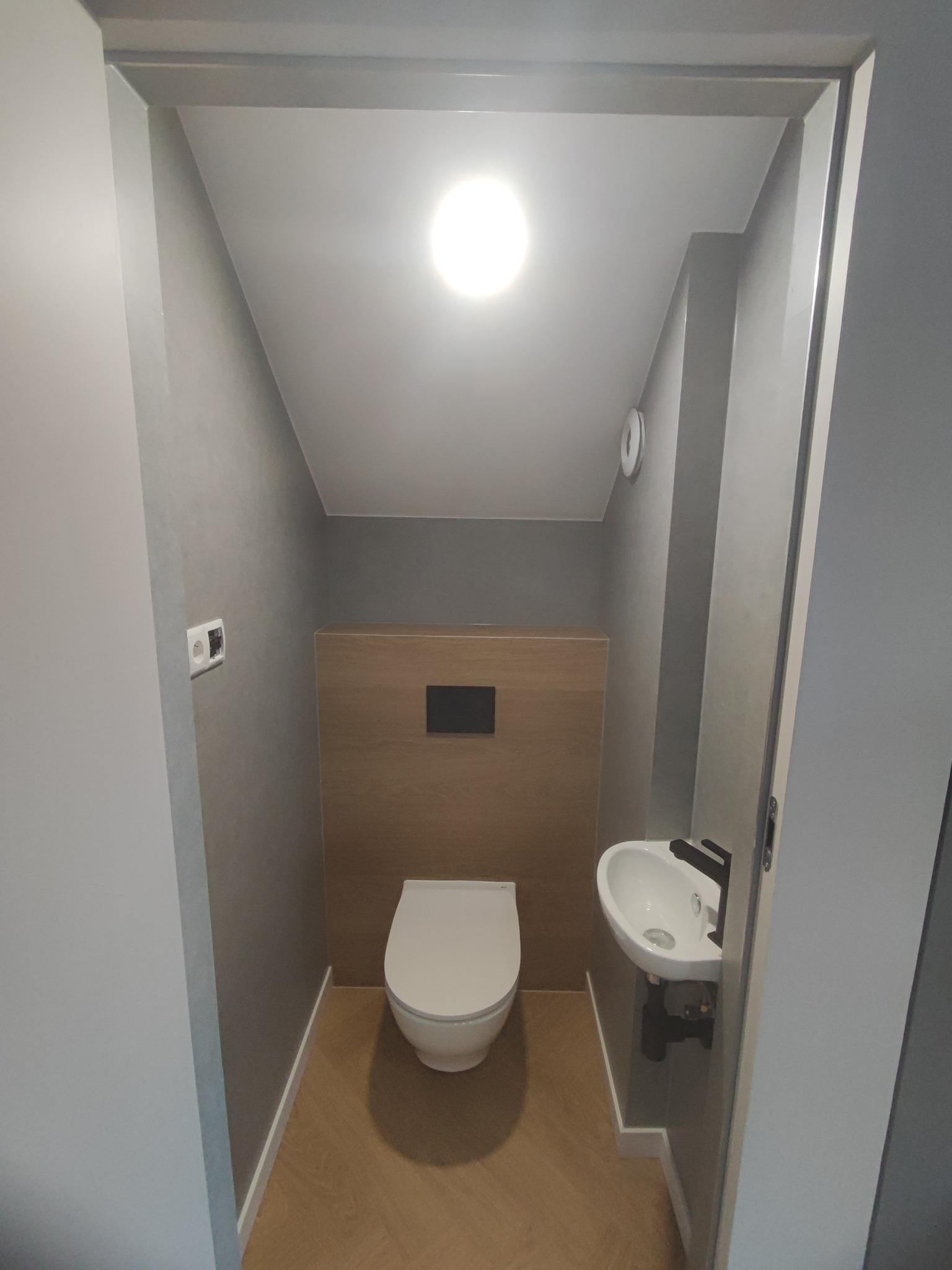 Nowoczesna, mała łazienka z toaletą podwieszaną, umywalką nablatową i drewnianą zabudową ściany, wykończona w szarościach i drewnie, oświetlona centralnym punktem świetlnym.