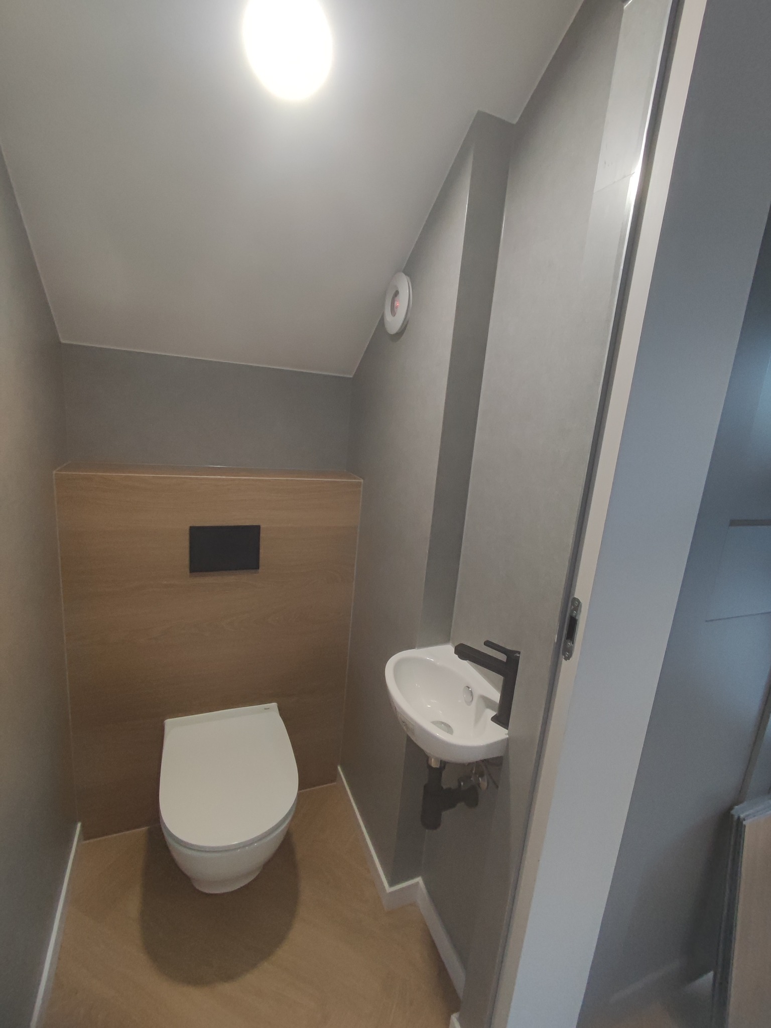 Nowoczesna, mała łazienka z wiszącą toaletą, umywalką z czarną armaturą, szarą ścianą i drewnopodobną zabudową oraz podłogą.