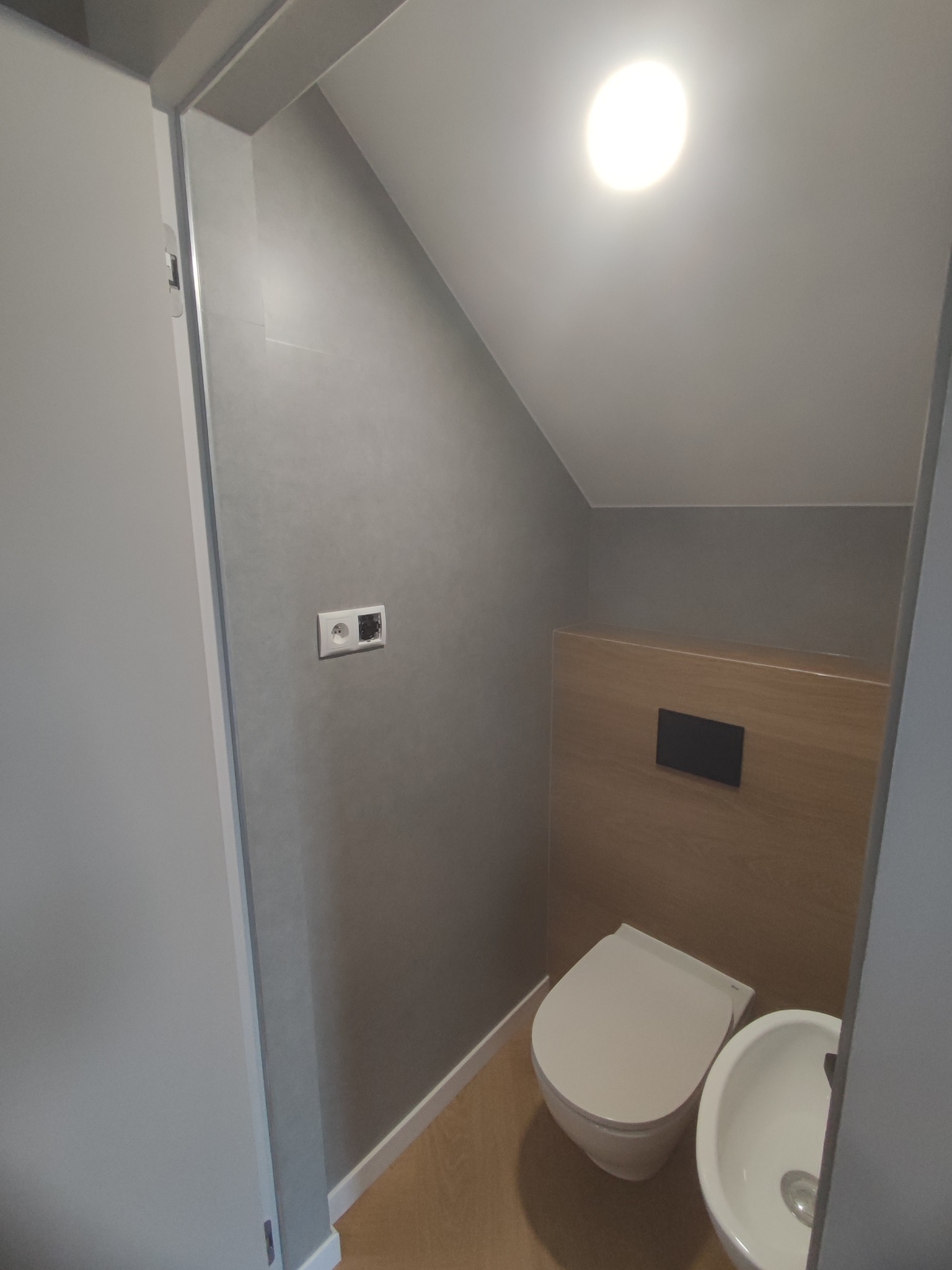 Nowoczesna, minimalistyczna łazienka na poddaszu z toaletą podwieszaną i umywalką, szare ściany, drewniana zabudowa, widoczny fragment gniazdka elektrycznego bez osprzętu.