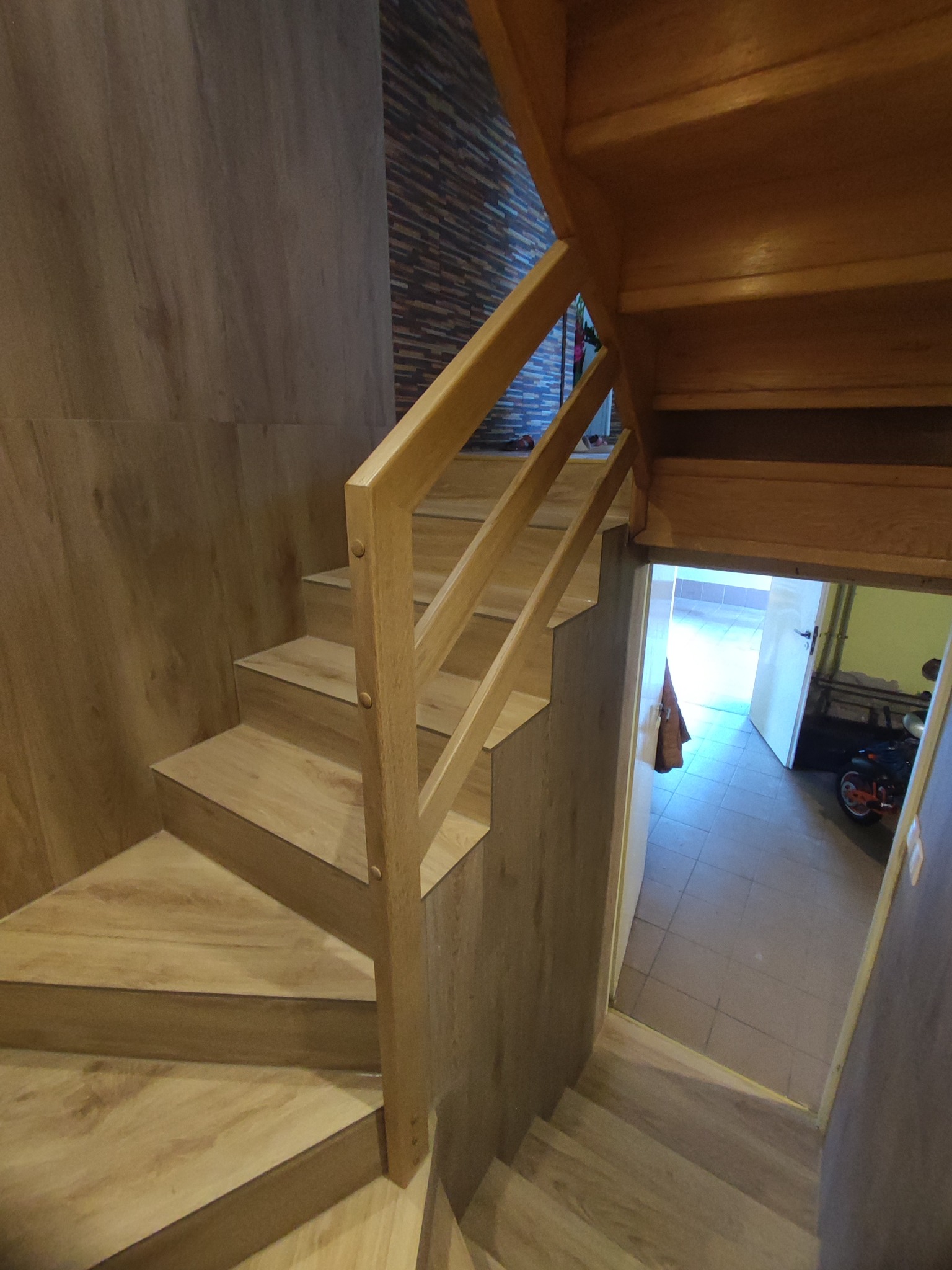 Drewniane schody z balustradą w wykończonym wnętrzu, z widoczną okładziną ścienną imitującą drewno i fragmentem pomieszczenia za otwartymi drzwiami.