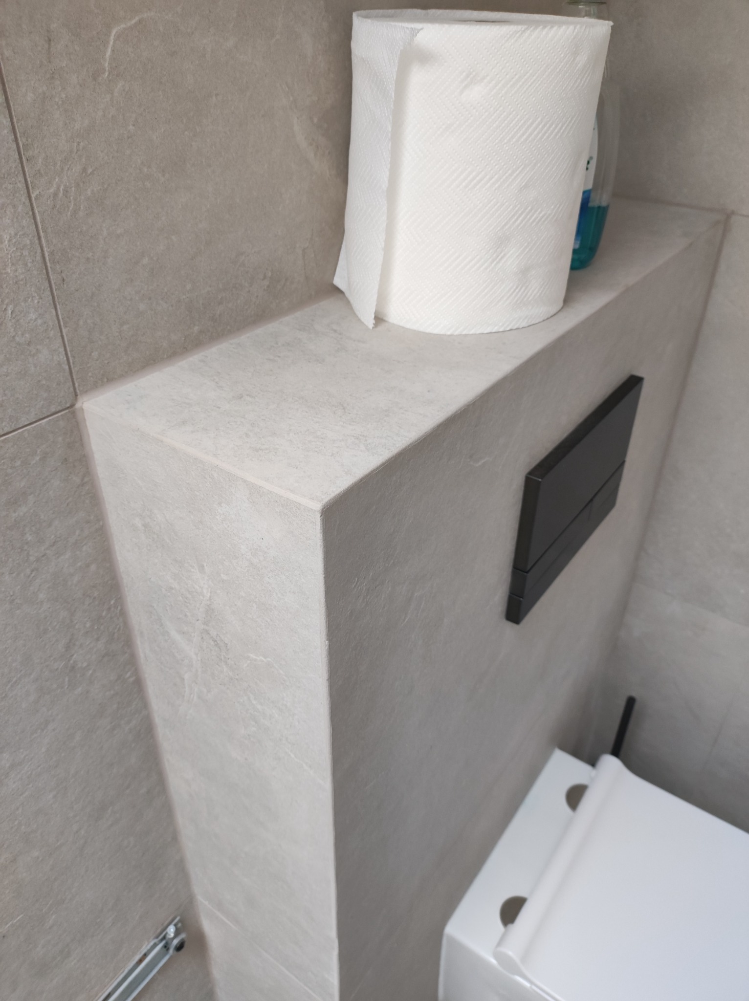 Szara, wykafelkowana zabudowa stelaża WC z czarnym przyciskiem spłukującym, rolką papieru toaletowego i butelką płynu na górze. Widoczny fragment muszli toaletowej.