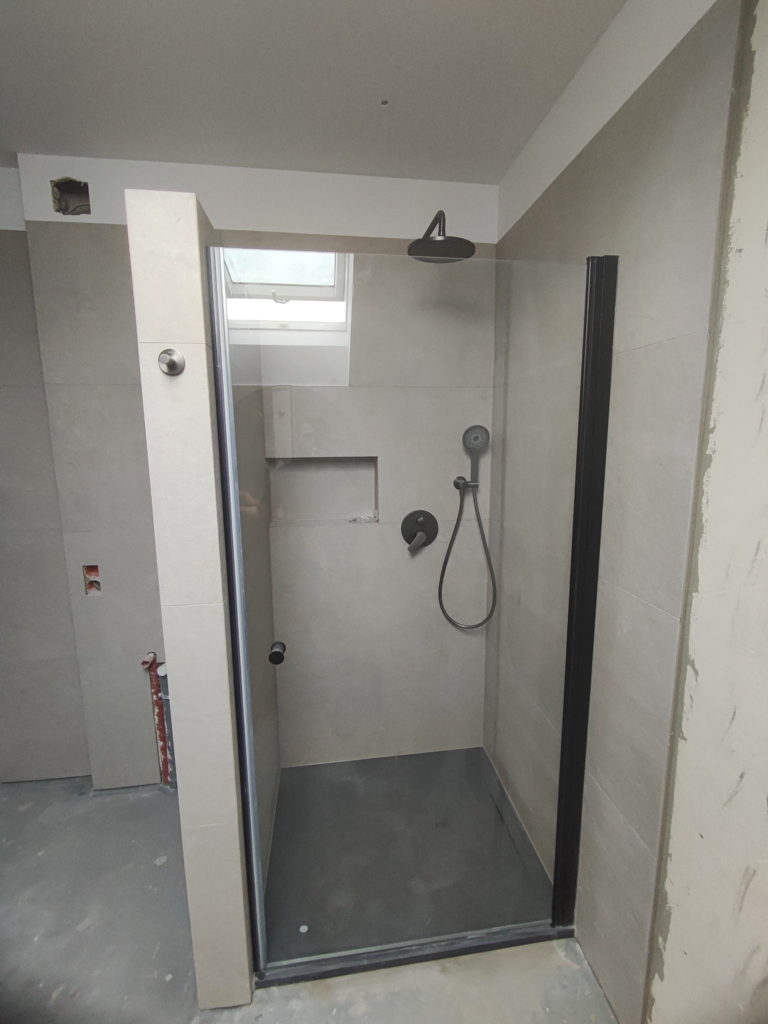 Nowoczesna kabina prysznicowa z czarnymi profilami i deszczownicą, szare płytki na ścianach, widoczne niedokończone prace wykończeniowe.