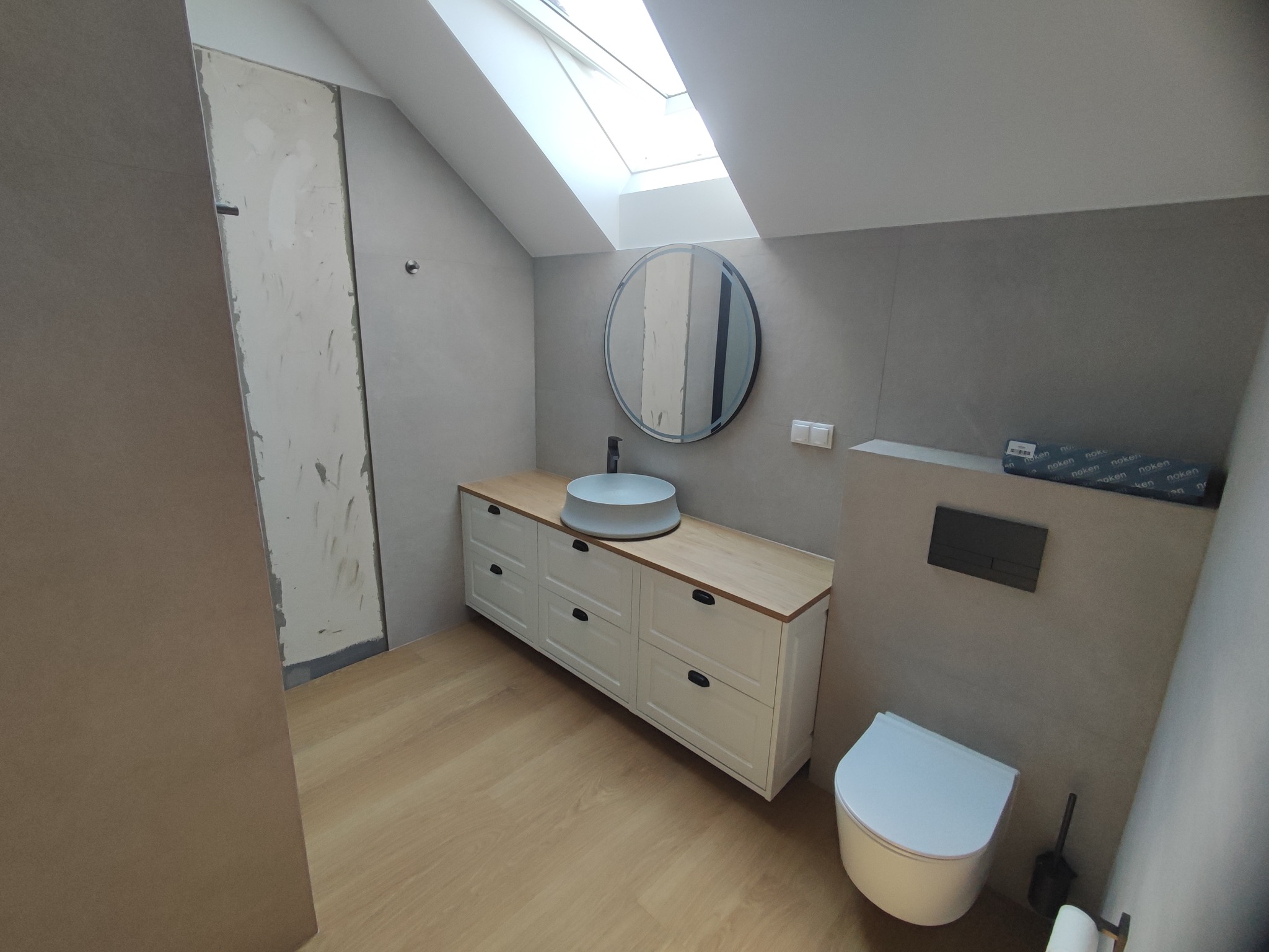 Nowoczesna łazienka w Porąbce Iwkowska z umywalką nablatową, okrągłym lustrem i podwieszaną toaletą, częściowo wykończona, z widoczną surową ścianą przed wykończeniem.