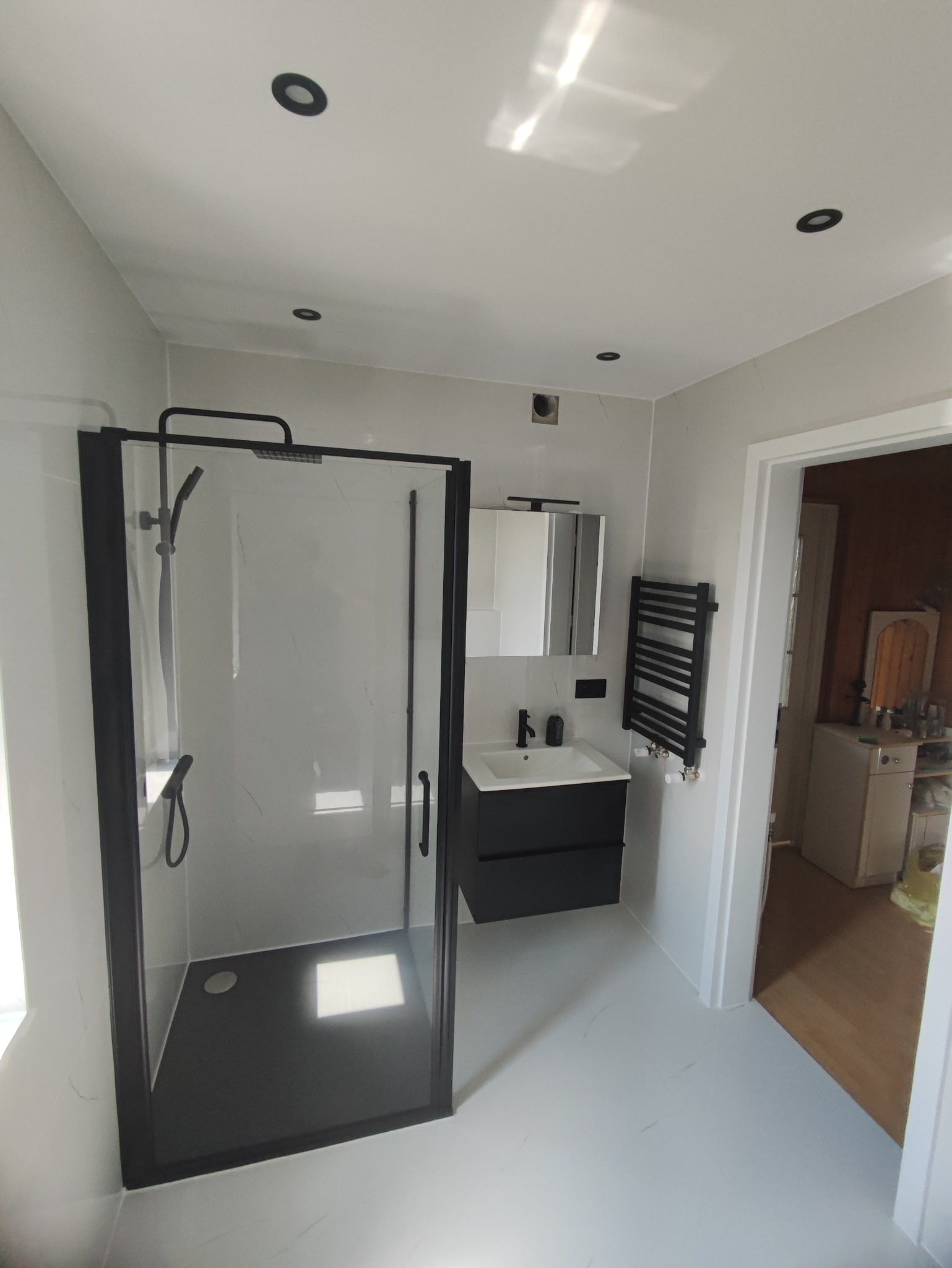 Nowoczesna łazienka z czarną kabiną prysznicową typu walk-in, umywalką z szafką w tym samym kolorze oraz grzejnikiem drabinkowym, całość utrzymana w jasnej kolorystyce z akcentami imitacji marmuru...