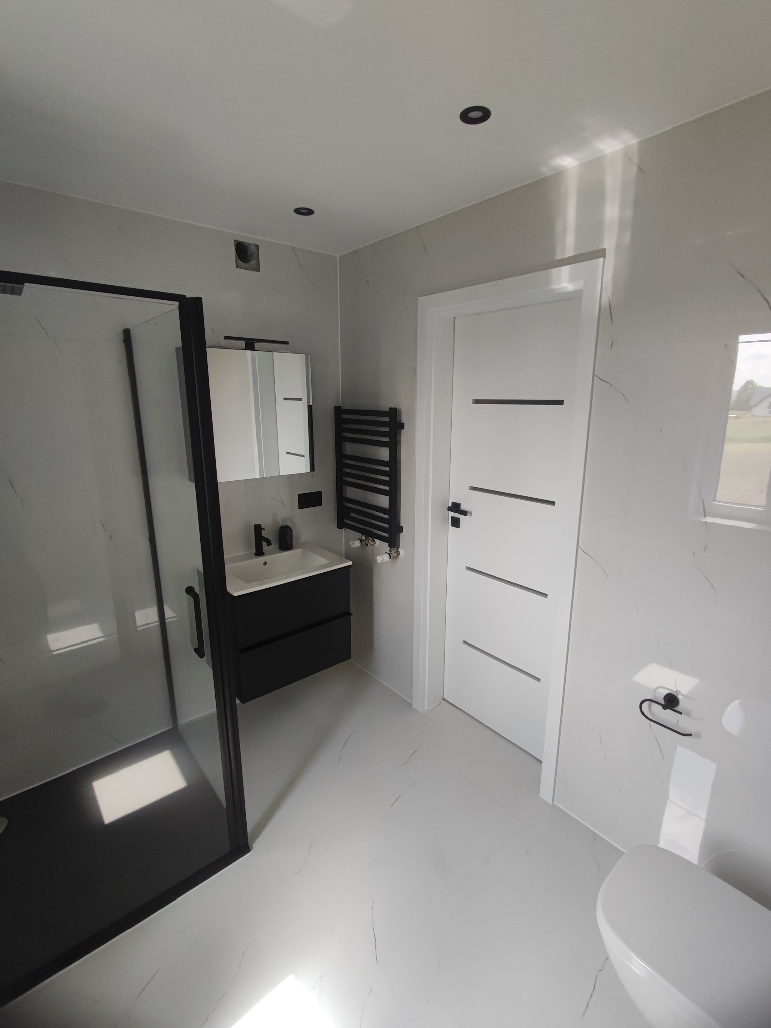 Nowoczesna łazienka w bieli i czerni: kabina prysznicowa z czarną ramą, wisząca szafka z umywalką, grzejnik drabinkowy i białe płytki imitujące marmur.