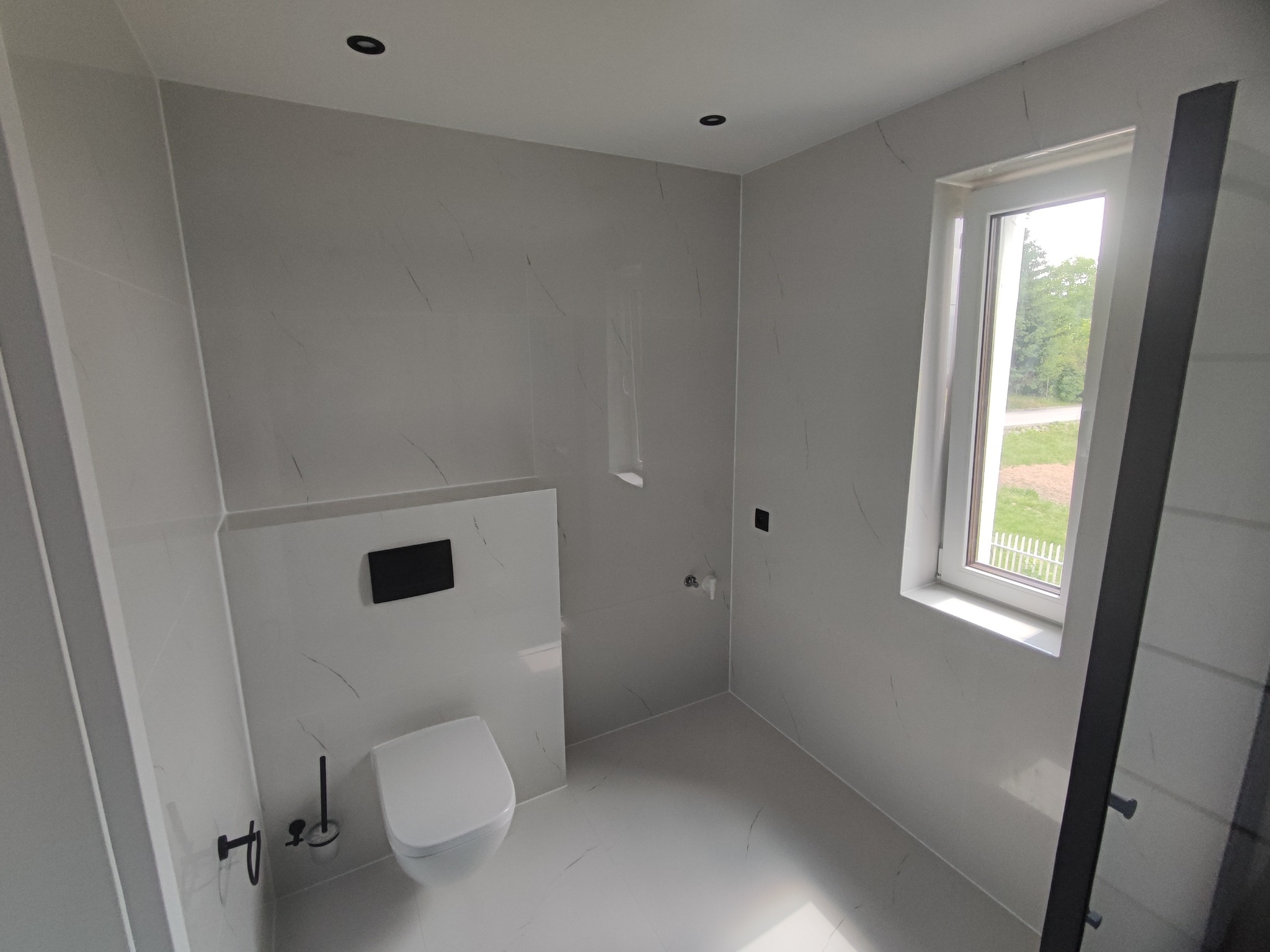 Nowoczesna łazienka z białymi płytkami imitującymi marmur, wisząca toaleta z czarnym przyciskiem spłukiwania, czarne akcesoria i okno z widokiem na zieleń.