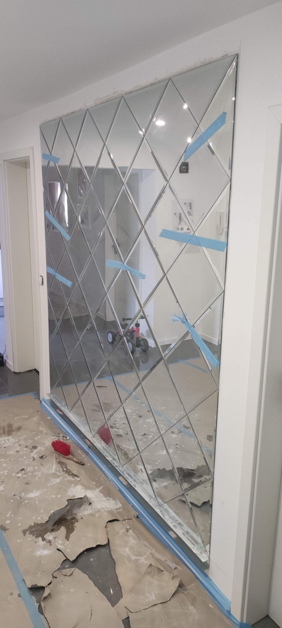 Ściana pokryta dekoracyjnymi lustrami w romby, zabezpieczona niebieską taśmą malarską, widoczne ślady prac wykończeniowych na podłodze i w tle, odblask pokoju w lustrach.