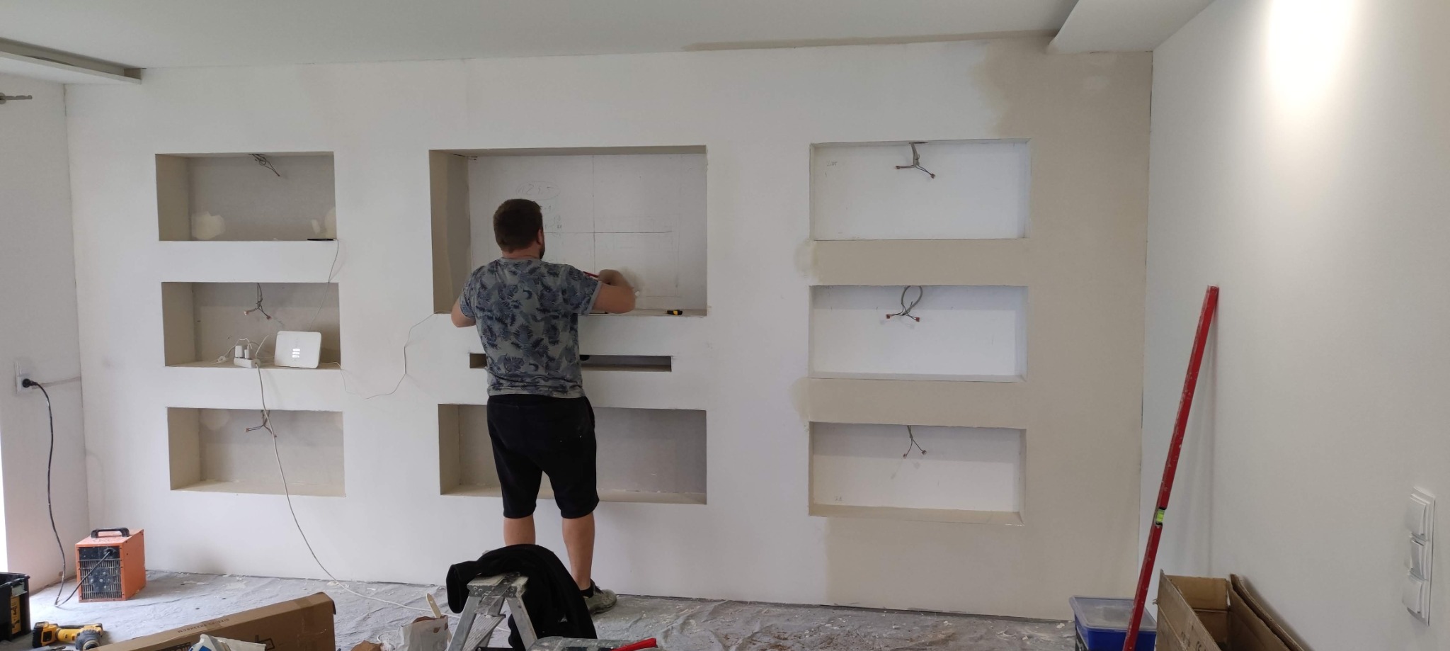 Mężczyzna w koszulce w kwiaty, stojący na drabinie, zaznacza ołówkiem linie na ścianie z wnękami w trakcie prac wykończeniowych, widoczne narzędzia i sprzęt budowlany.
