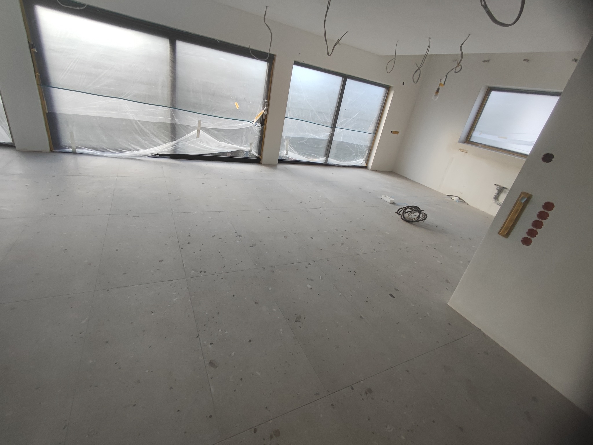 Wnętrze w trakcie remontu: szare płytki podłogowe, okna zabezpieczone folią malarską, widoczne przewody elektryczne zwisające z sufitu, przygotowanie do malowania.