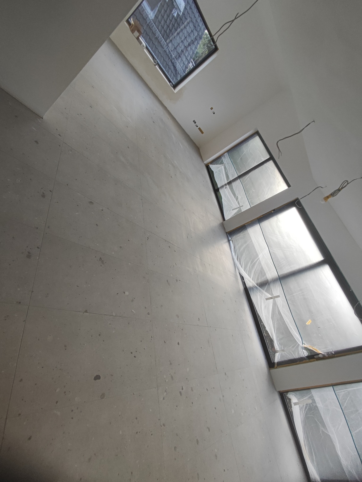Wnętrze w trakcie prac wykończeniowych, widoczne szare płytki na podłodze i ścianie, okna zabezpieczone folią malarską, oraz wystające kable elektryczne.