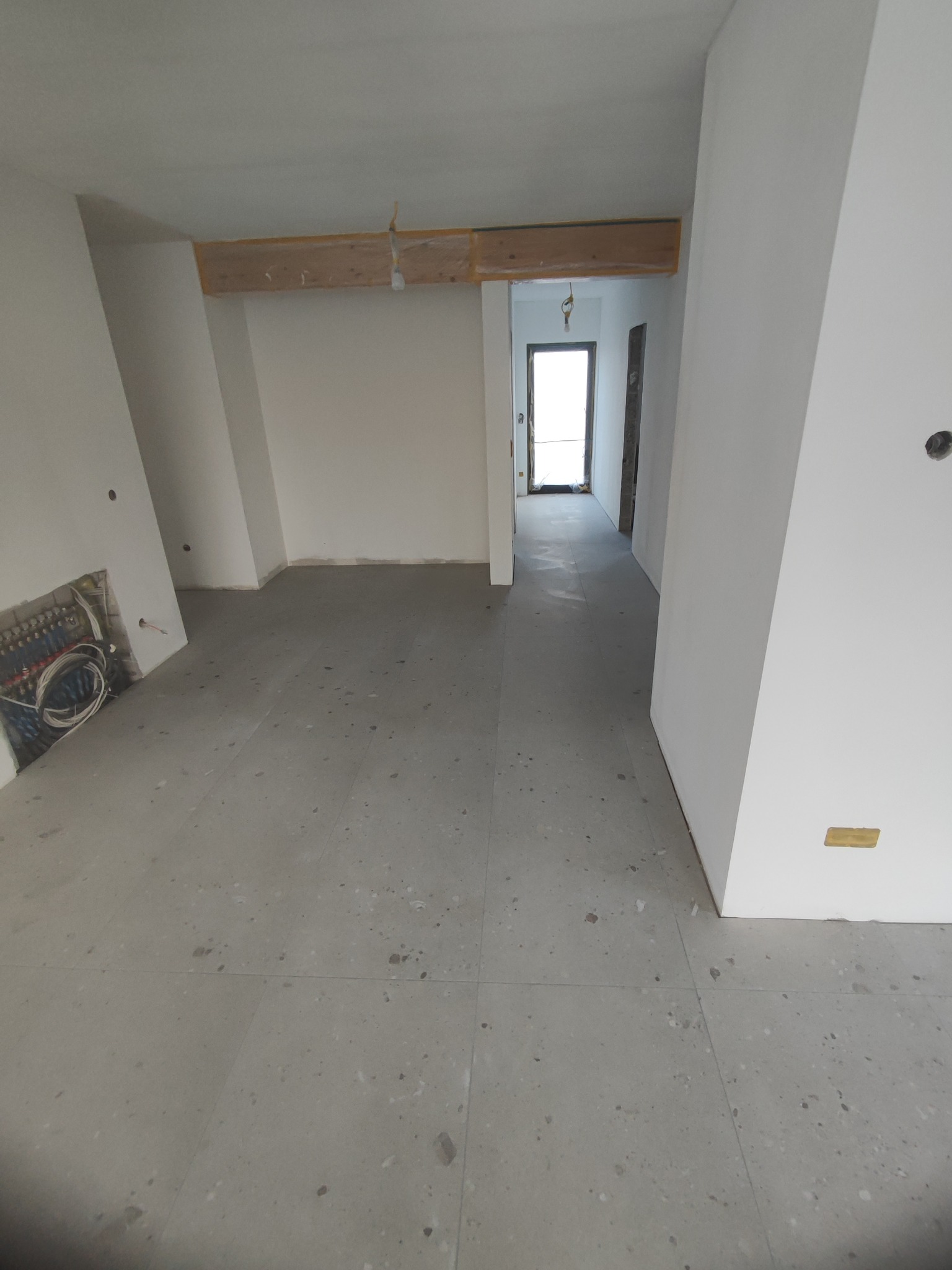 Surowe wnętrze w trakcie prac wykończeniowych: szare płytki podłogowe, białe ściany z widocznymi instalacjami, drewniane elementy konstrukcyjne pod sufitem, widok na korytarz.