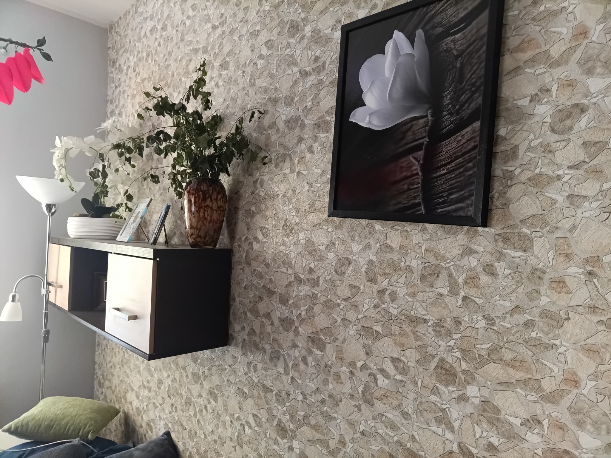 Półka wisząca w salonie z dekoracjami, na ścianie tapeta imitująca kamień i obraz z kwiatem.