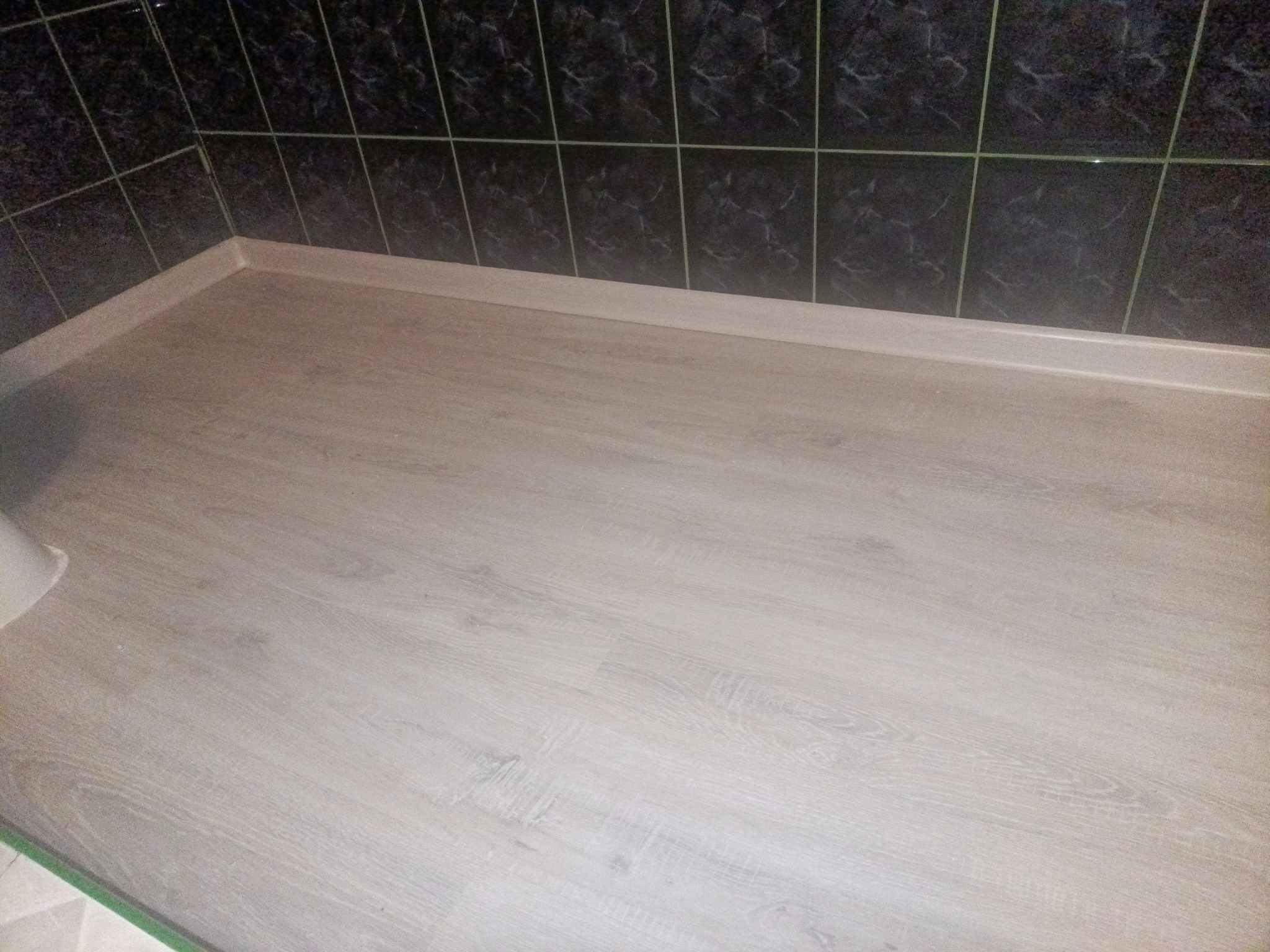 Jasne panele podłogowe ułożone w łazience w Nowym Miasteczku, widoczne ciemne płytki na ścianie i biała listwa przypodłogowa.