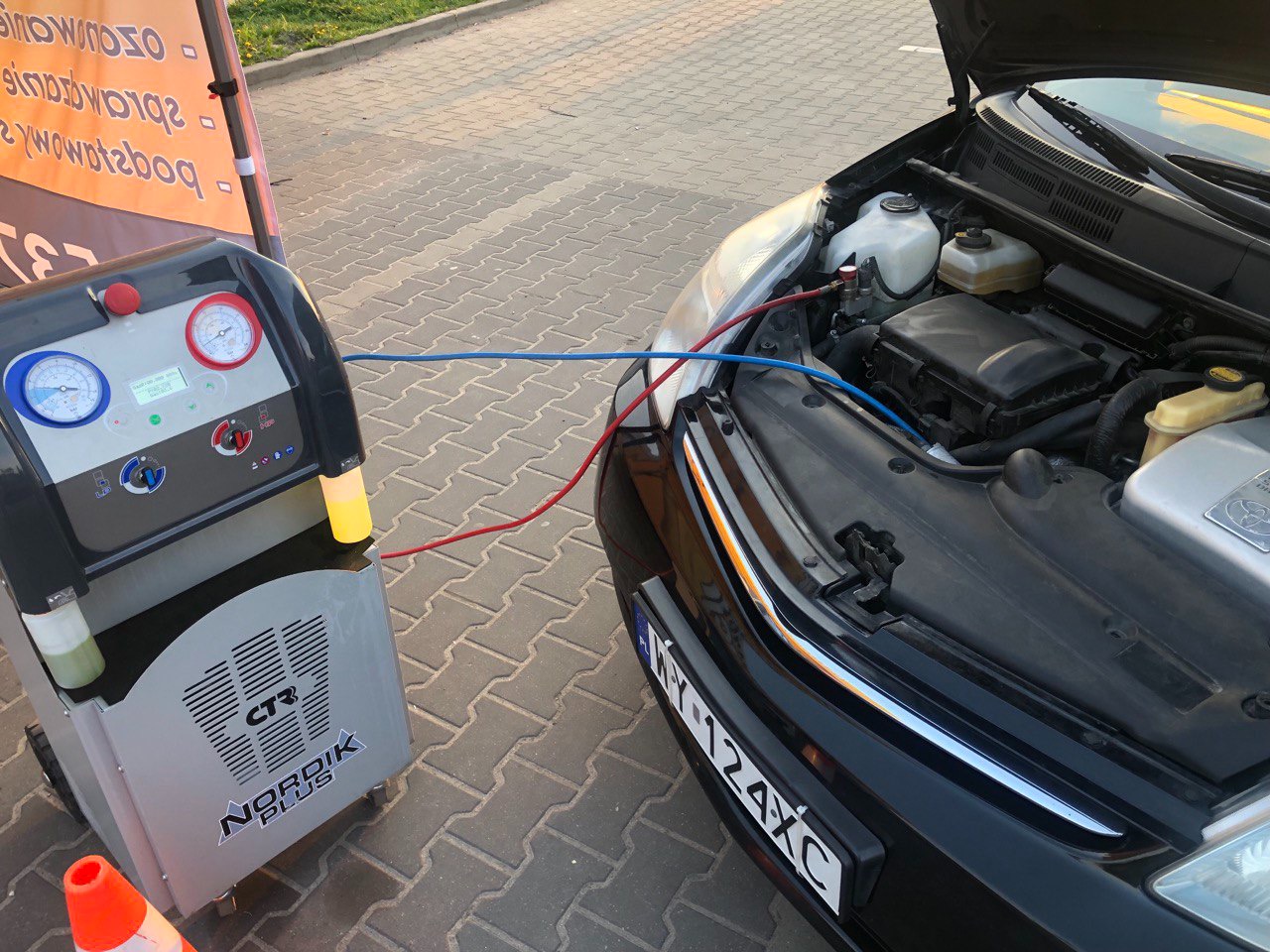 Czarny samochód z otwartą maską podłączony przewodami do szarego urządzenia diagnostycznego z manometrami i wyświetlaczem; widoczny silnik i elementy instalacji klimatyzacji.