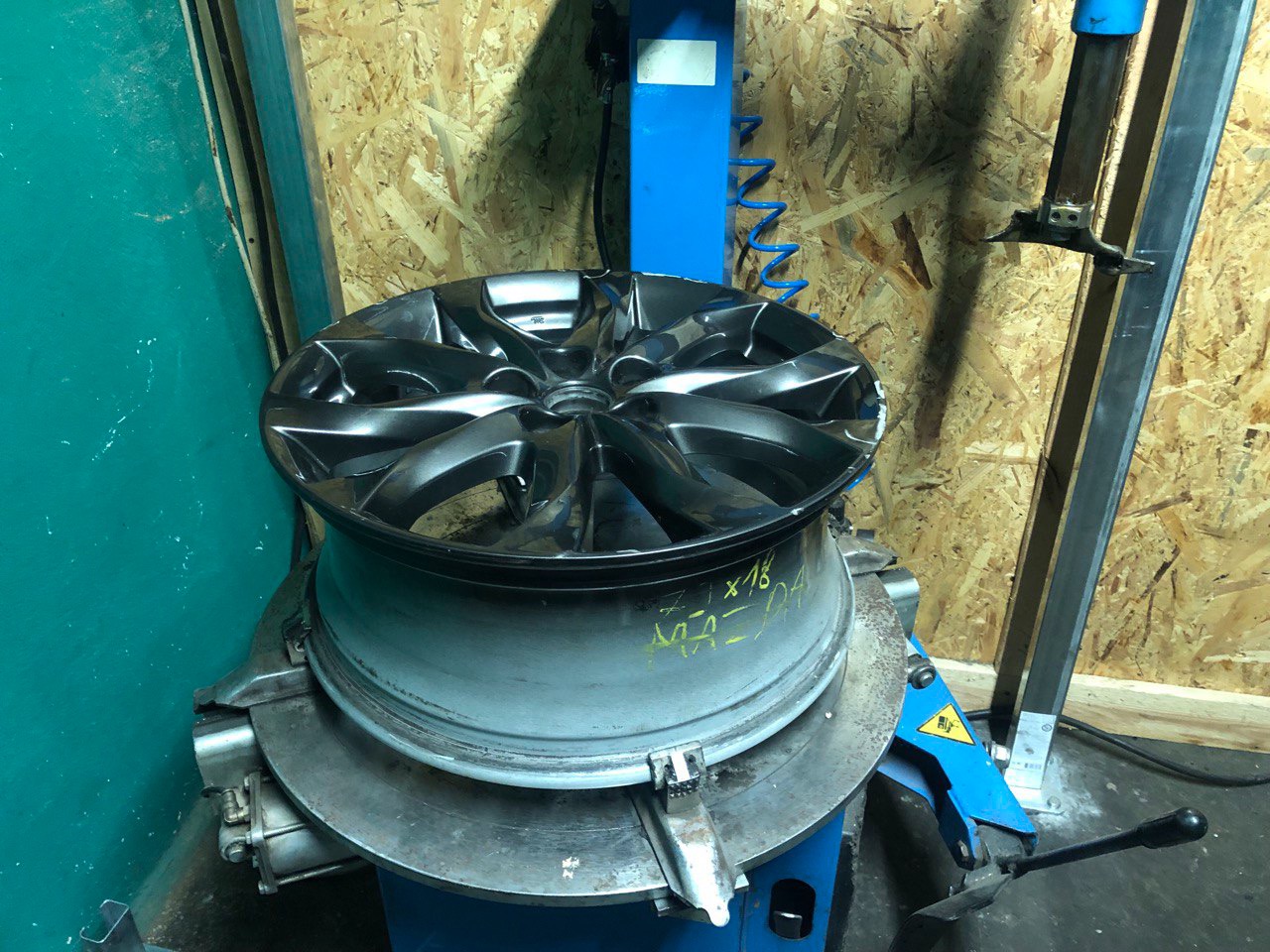 Czarna alufelga Mazda na montażownicy w warsztacie wulkanizacyjnym, z widocznymi żółtymi napisami i oznaczeniami na feldze.