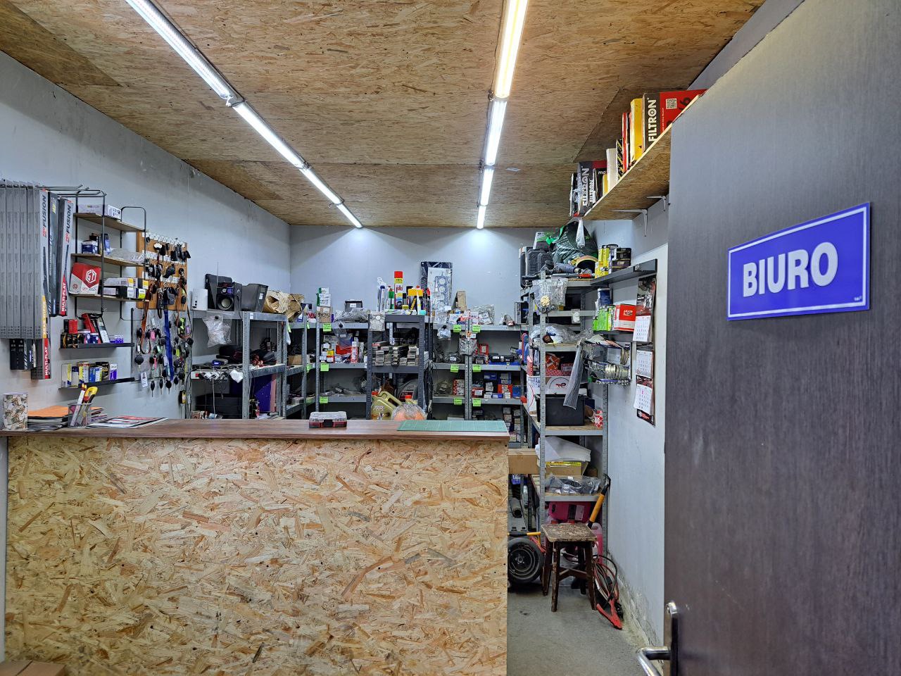 Wnętrze biura warsztatu samochodowego z ladą z płyty OSB, regałami pełnymi części i akcesoriów oraz niebieskim znakiem 'Biuro' na drzwiach.
