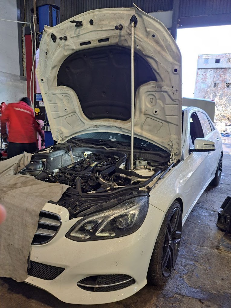 Biały Mercedes z otwartą maską silnika w warsztacie, zabezpieczony szarą płachtą, widoczny mechanik w czerwonej kurtce w tle.