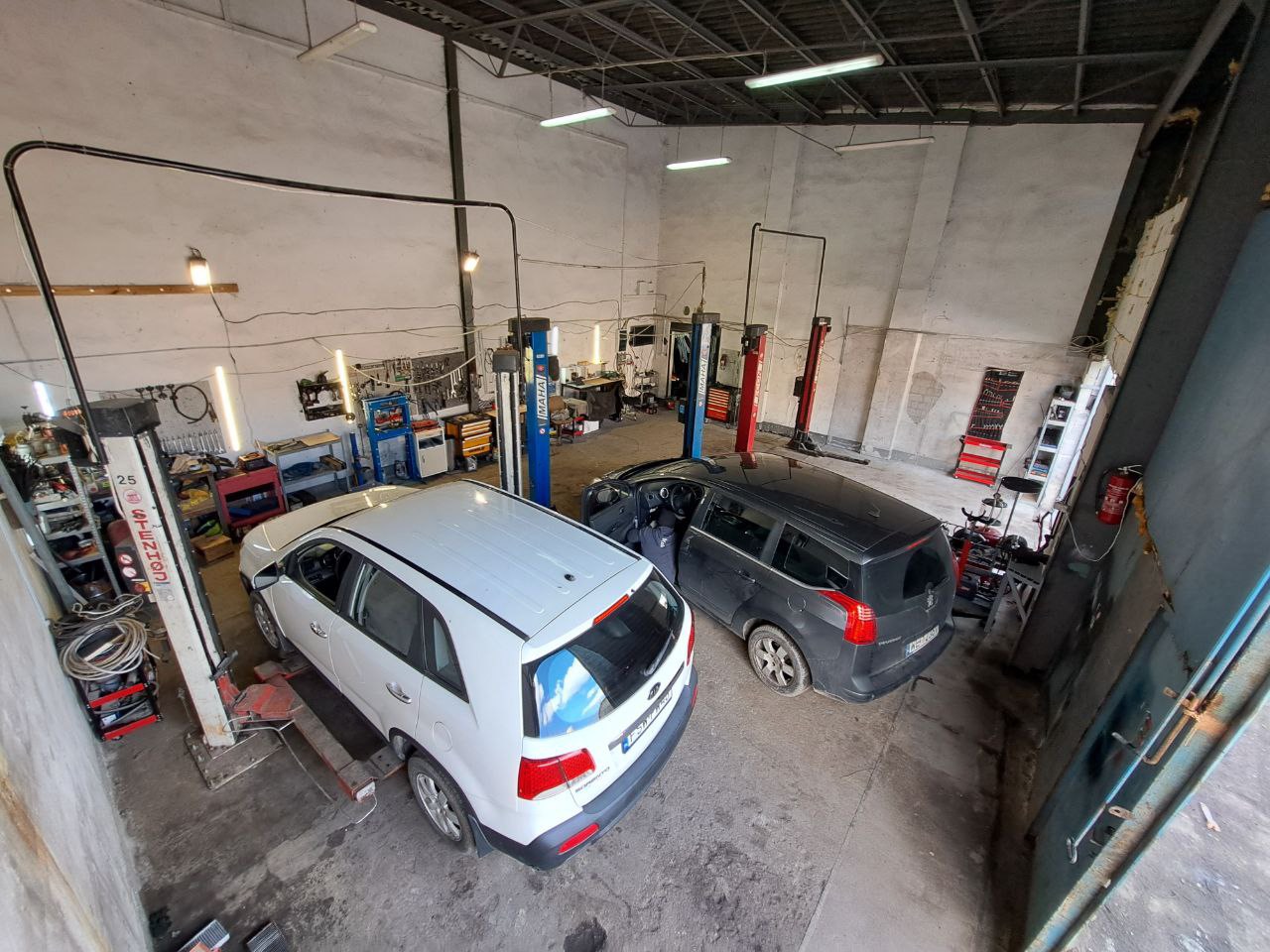 Wnętrze warsztatu samochodowego z dwoma samochodami na podnośnikach, biały SUV i ciemny kombi, widok z góry na stanowiska pracy i narzędzia.