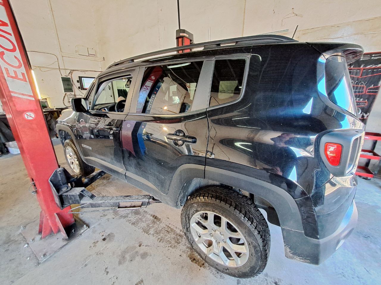 Czarny SUV na podnośniku w warsztacie, widoczne ślady użytkowania na karoserii i oponach, czerwony podnośnik kolumnowy, narzędzia na ścianie w tle.