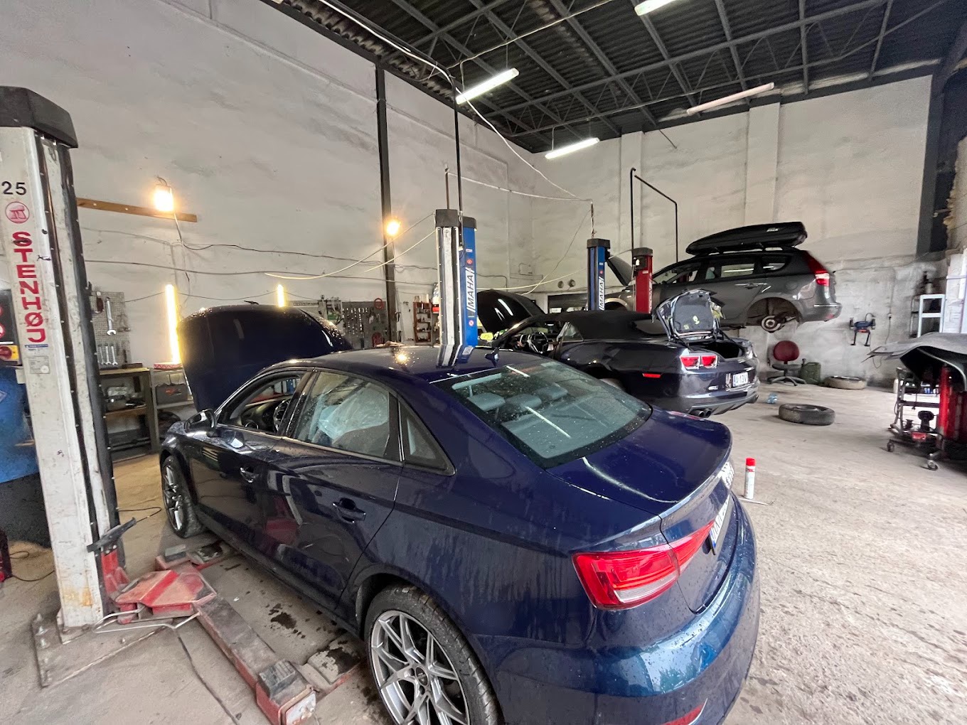 Wnętrze warsztatu samochodowego z podnośnikami, gdzie na jednym z nich stoi granatowy sedan Audi z otwartą maską, a w tle widoczne są inne pojazdy, w tym czarny kabriolet i szary SUV na podnośniku.