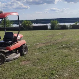 Czerwona kosiarka samojezdna na trawniku z widocznymi śladami koszenia, w tle jezioro i drzewa pod błękitnym niebem z chmurami. Słoneczny dzień.