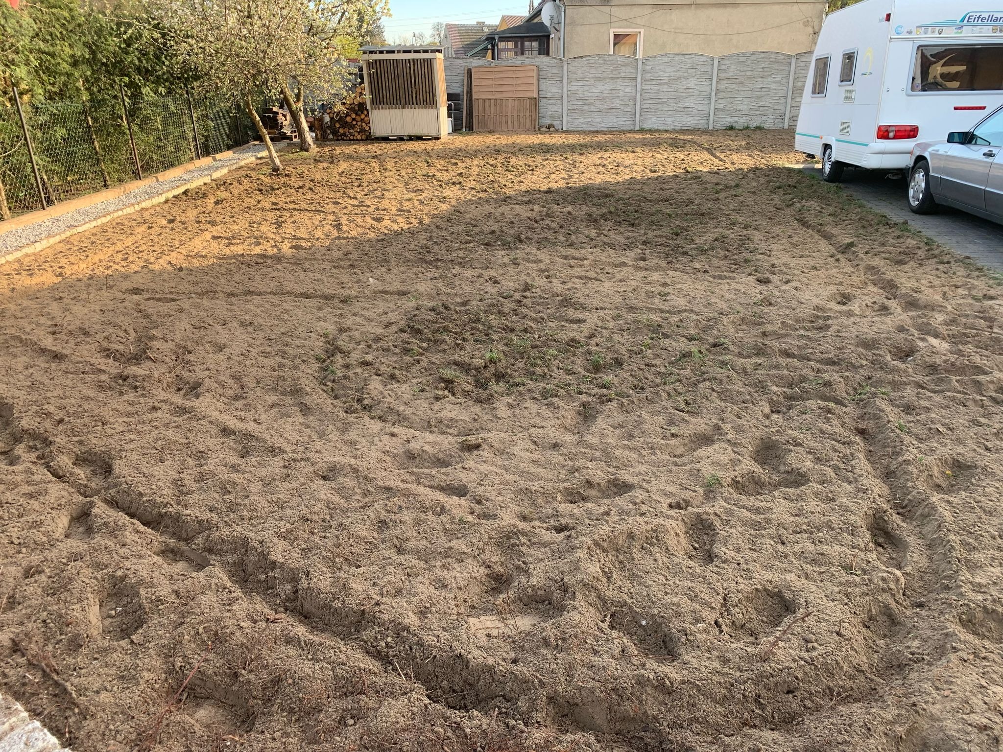 Przygotowany teren pod trawnik: wyrównana ziemia, ślady opon, widoczne budynki w tle. Prace ogrodnicze przed założeniem trawy.