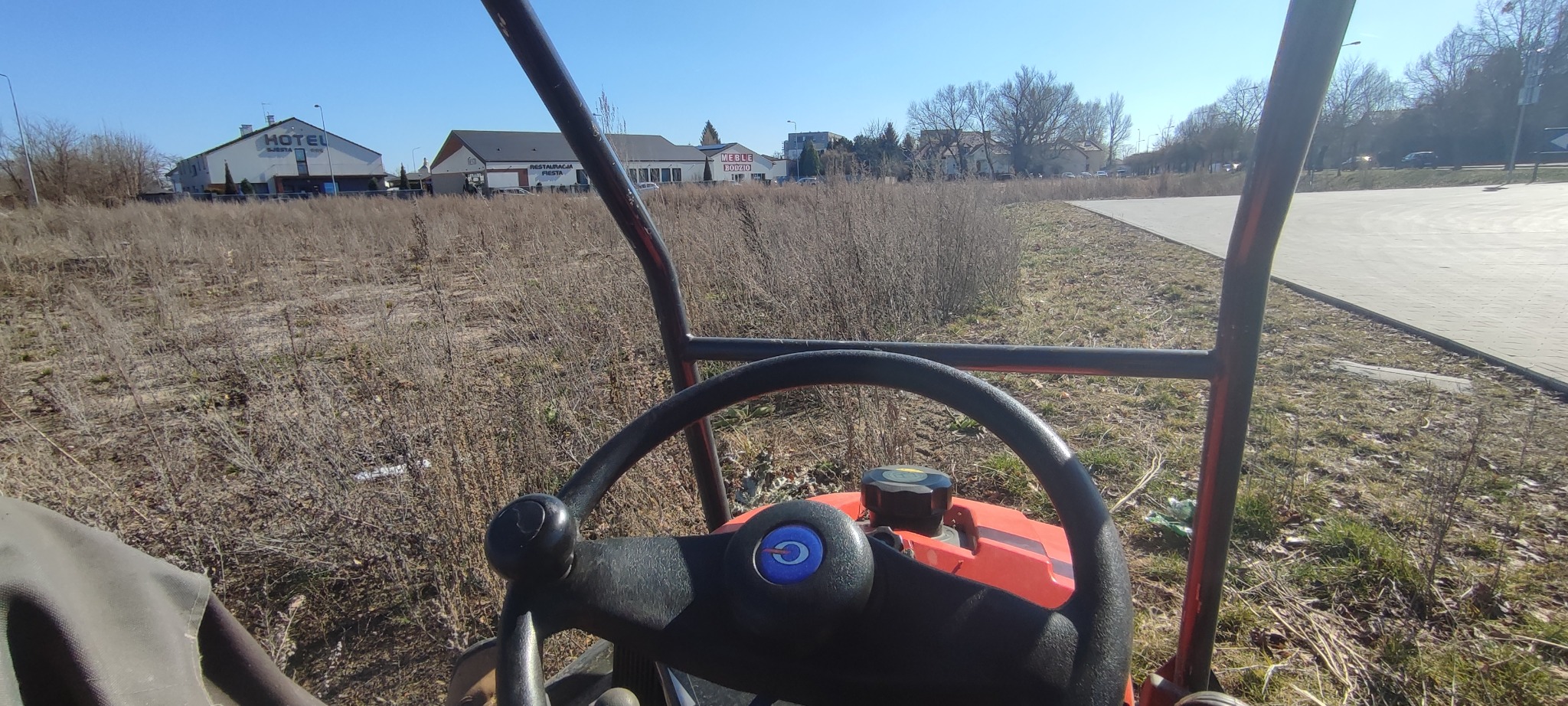 Widok z perspektywy operatora traktorka koszącego zaniedbane pole w Czerwieńsku. W tle budynki, w tym Hotel Sielsko Anielsko.