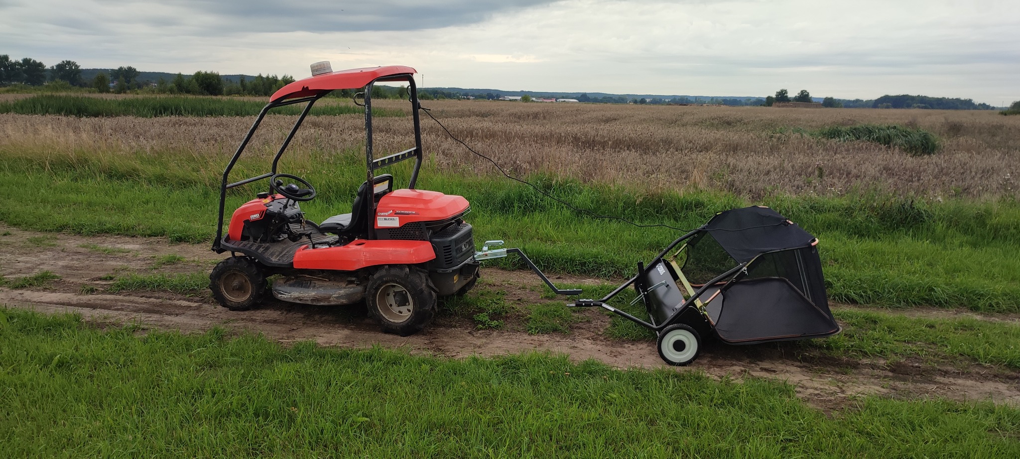 Czerwony traktorek ogrodowy z przyczepką na trawie, w tle pole i pochmurne niebo. Sprzęt gotowy do pracy w ogrodzie.