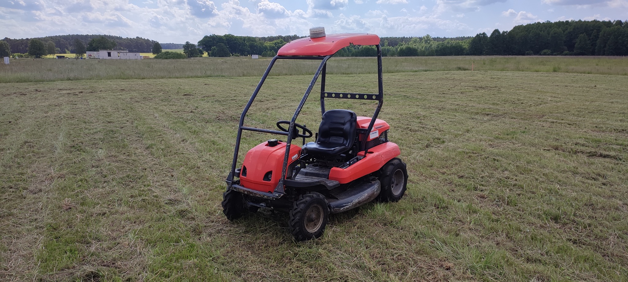 Czerwona kosiarka samojezdna na polu z częściowo skoszoną trawą. W tle widoczny las i budynki. Lekkie zachmurzenie.