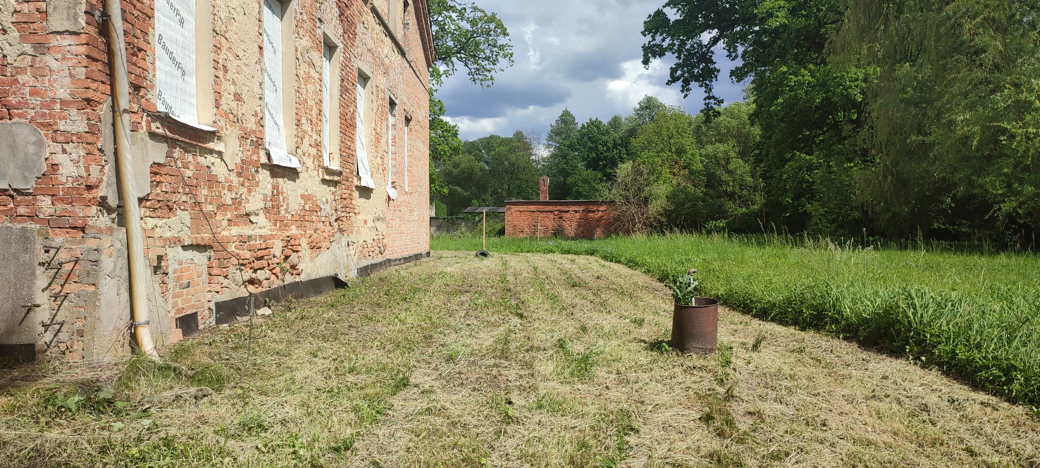 Trawa skoszona przy ceglanym budynku z oknami zakrytymi folią. W tle widoczna zieleń i budynek gospodarczy. Częściowo skoszona trawa.