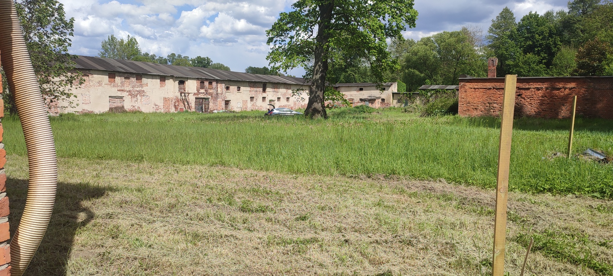 Rozległy teren z częściowo skoszoną trawą, budynki w tle. Duża, beżowa rura po lewej stronie. Widoczne drewniane słupki i samochód z otwartym bagażnikiem.