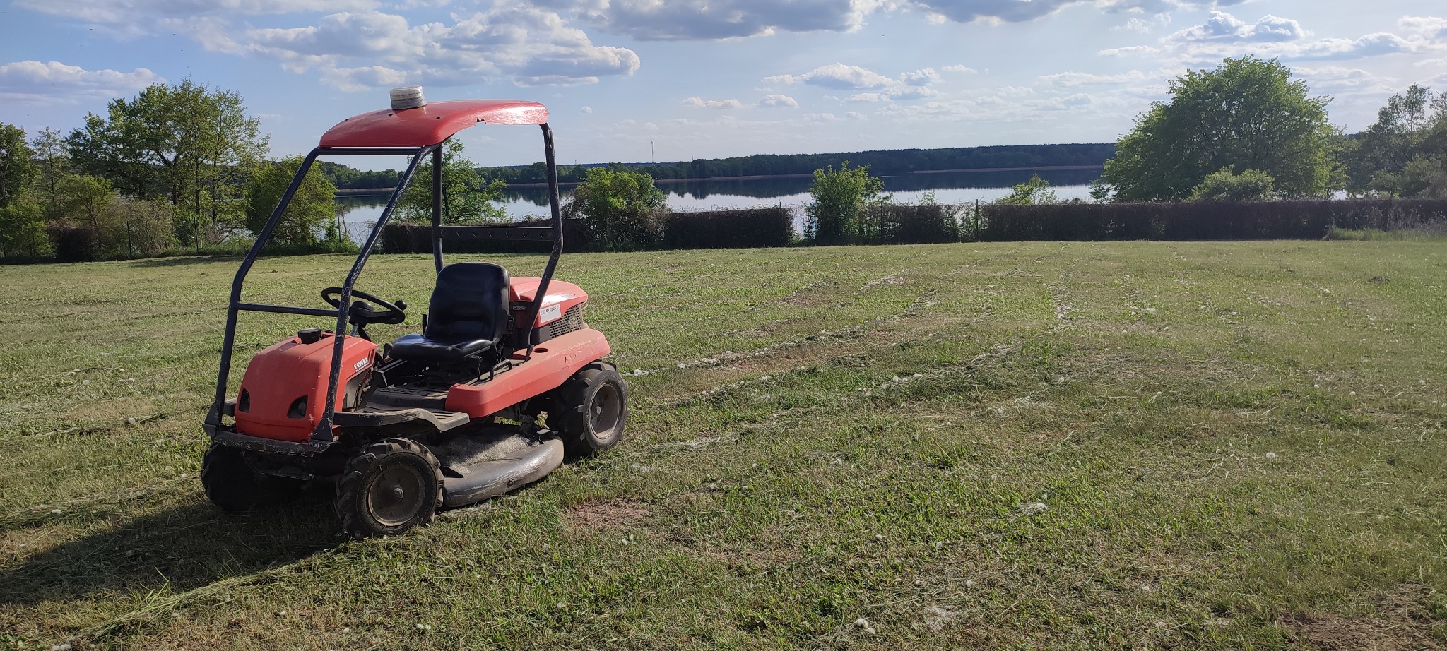 Czerwona kosiarka samojezdna na trawniku z widocznymi śladami koszenia, w tle jezioro i drzewa pod błękitnym niebem z chmurami. Słoneczny dzień.