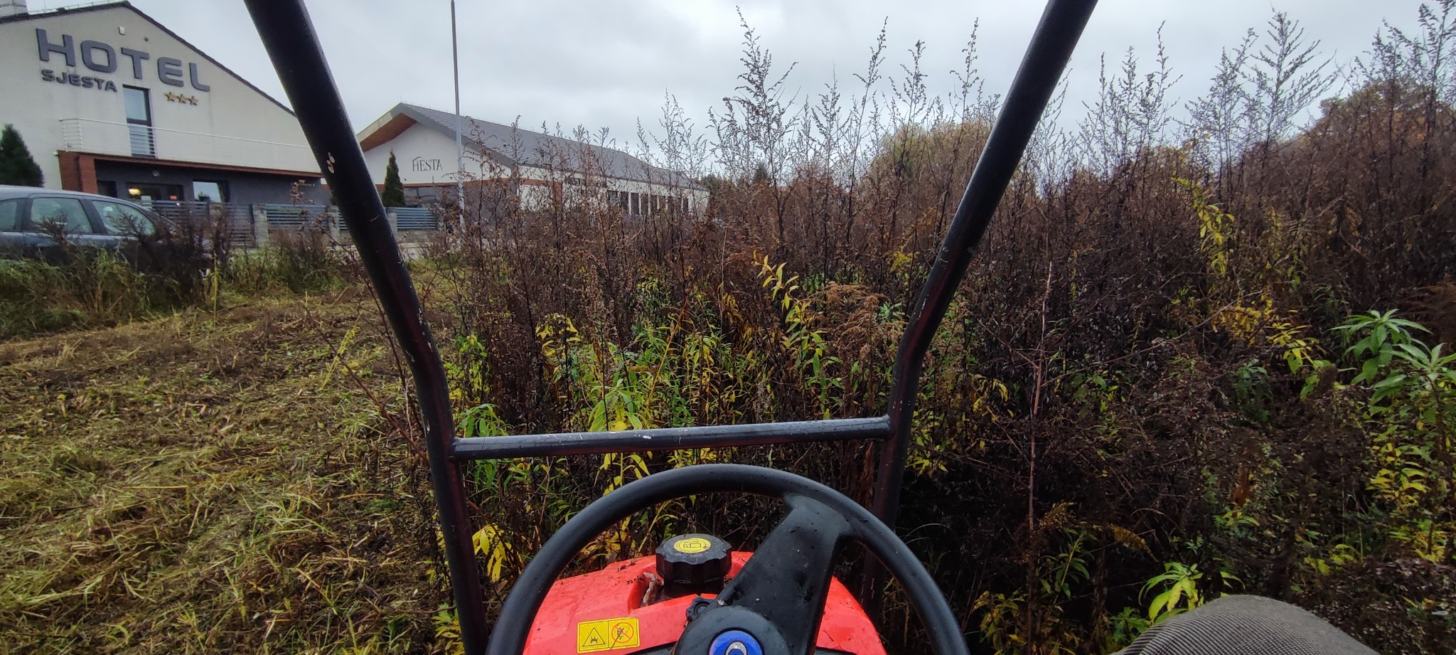 Widok z perspektywy operatora na teren zarośnięty wysoką trawą, częściowo skoszoną, z budynkami w tle. Widoczny fragment maszyny do koszenia.
