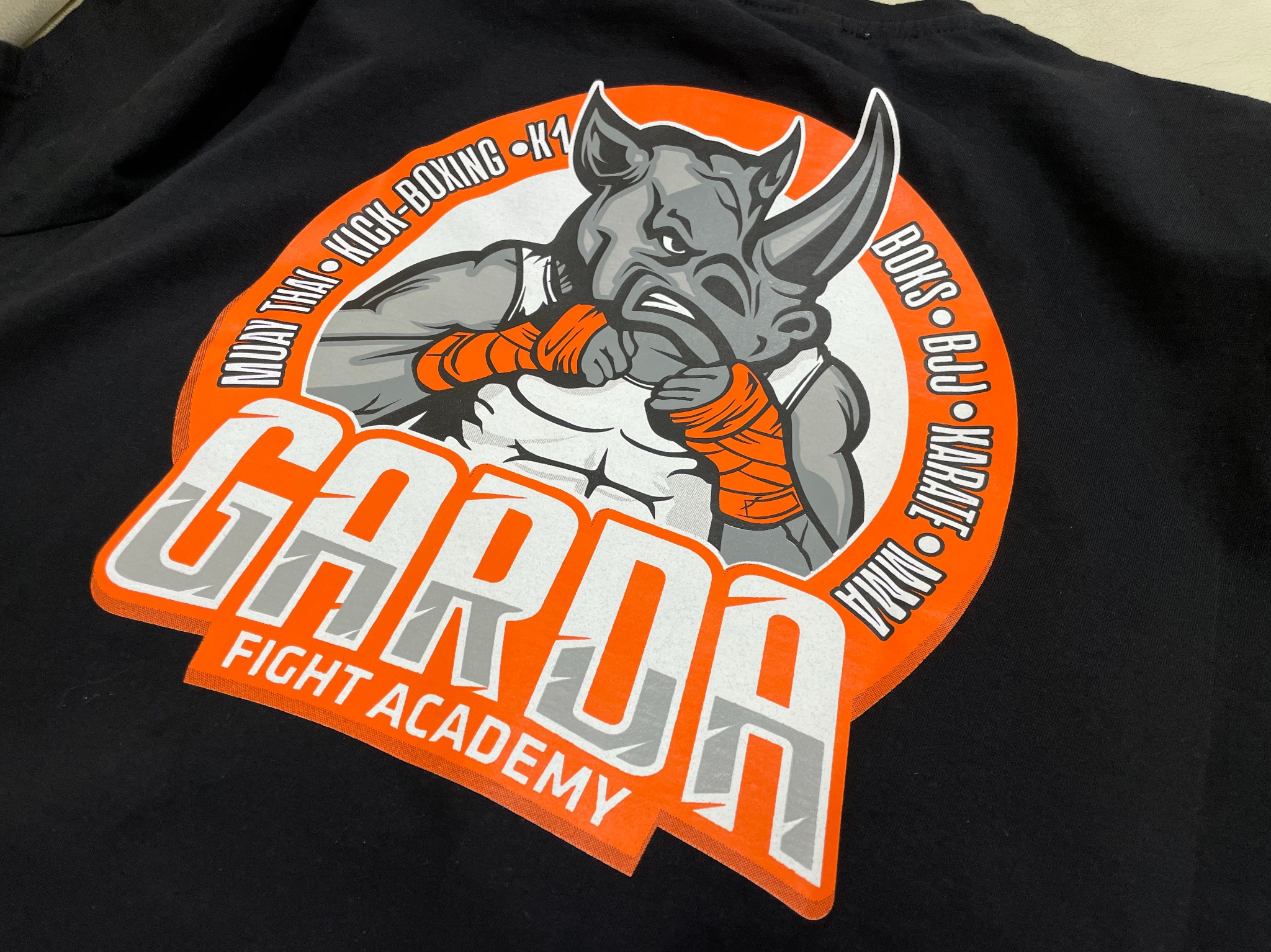 Czarna koszulka z nadrukiem logo 'GARDA FIGHT ACADEMY' przedstawiającym boksującego nosorożca, otoczonego nazwami sportów walki: Muay Thai, Kick-Boxing, K1, Boks, BJJ, Karate, MMA. Nadruk...