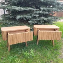 Dwie drewniane szafki nocne z otwartymi szufladami, ustawione na trawie przed iglastym drzewem.