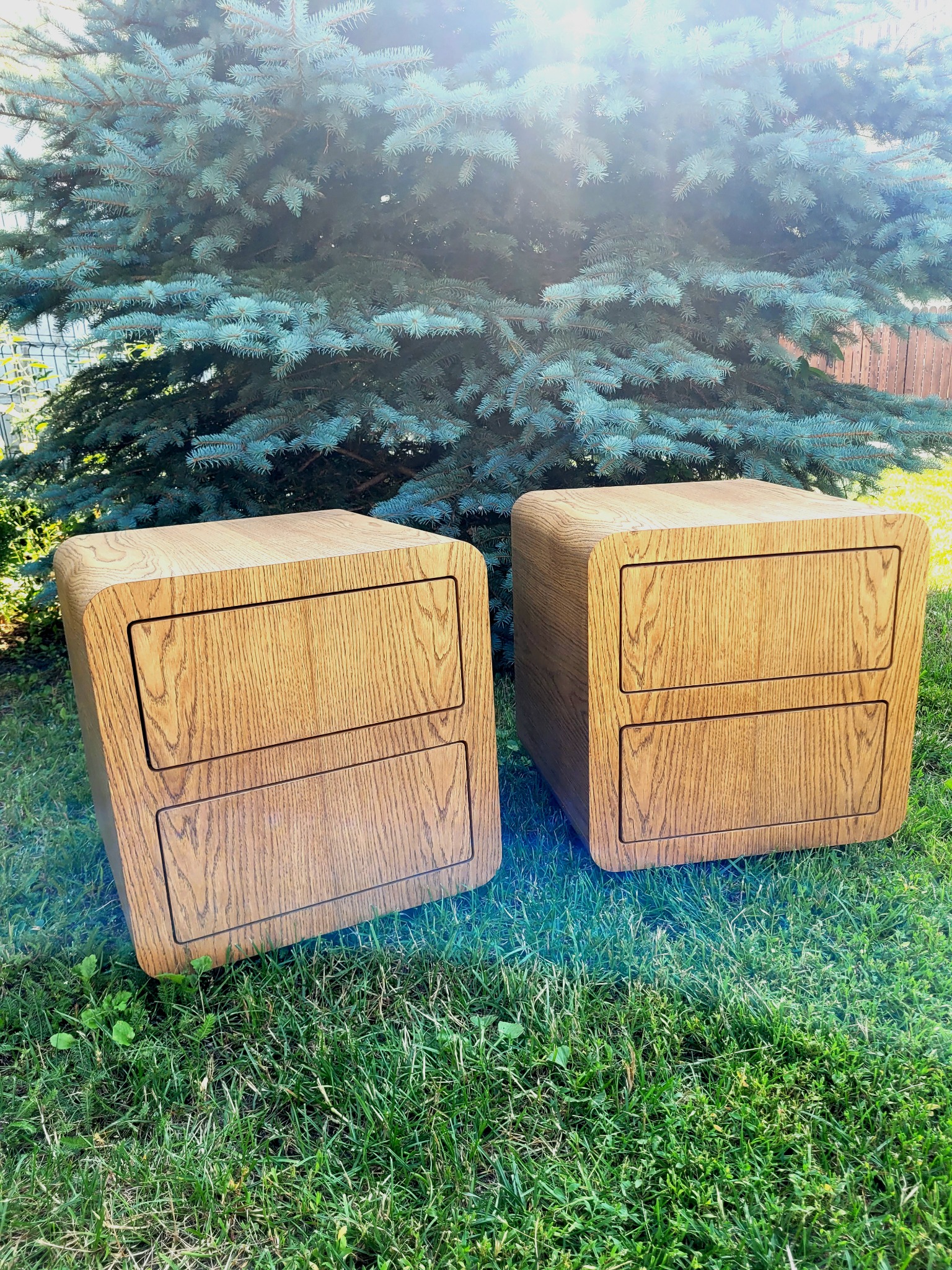 Dwie drewniane szafki nocne z dwiema szufladami, zaokrąglone krawędzie, ustawione na trawie przed świerkiem.