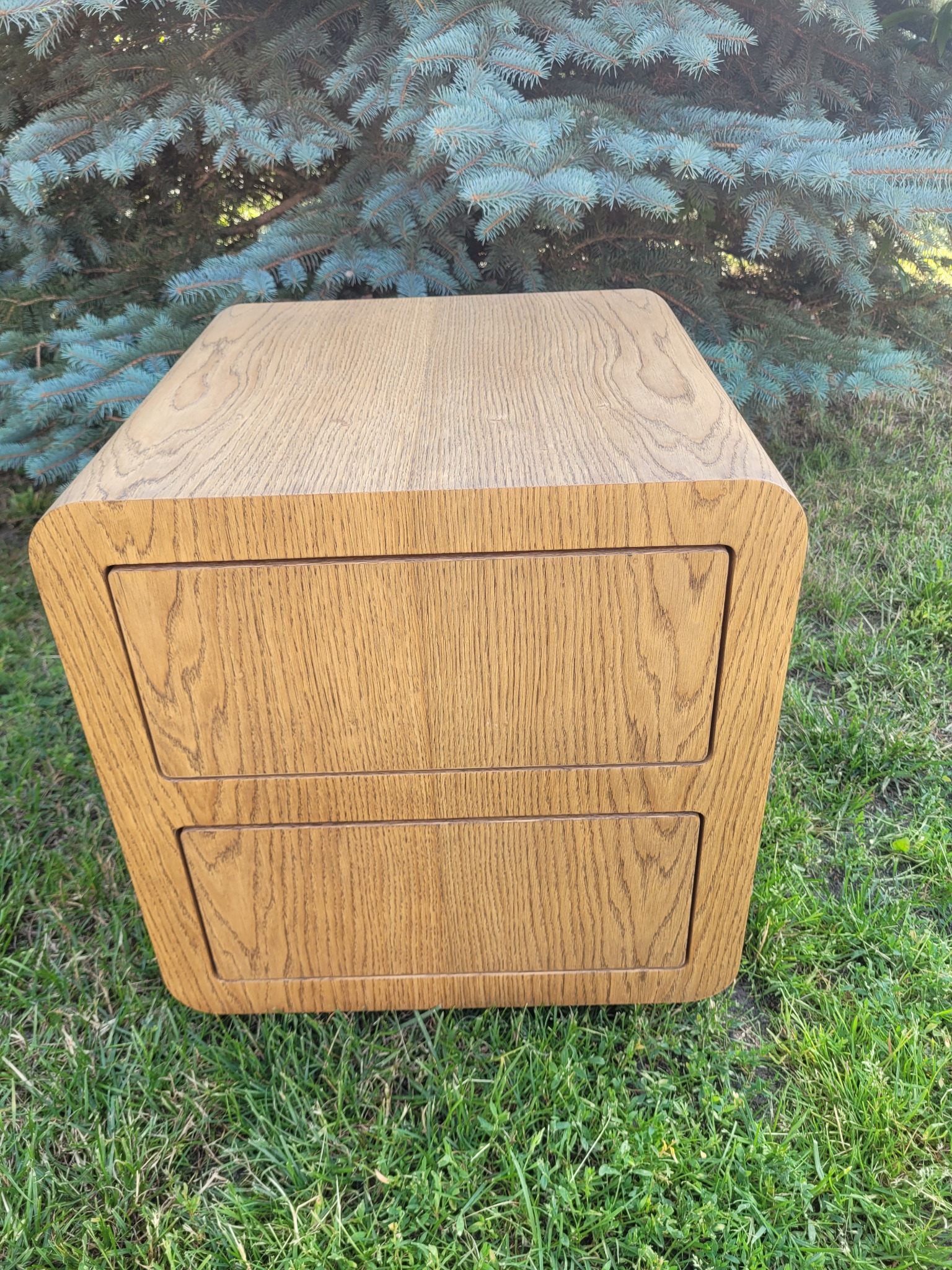 Dębowa komoda z dwiema szufladami, zaokrąglone krawędzie, na trawie, z iglastym drzewem w tle.