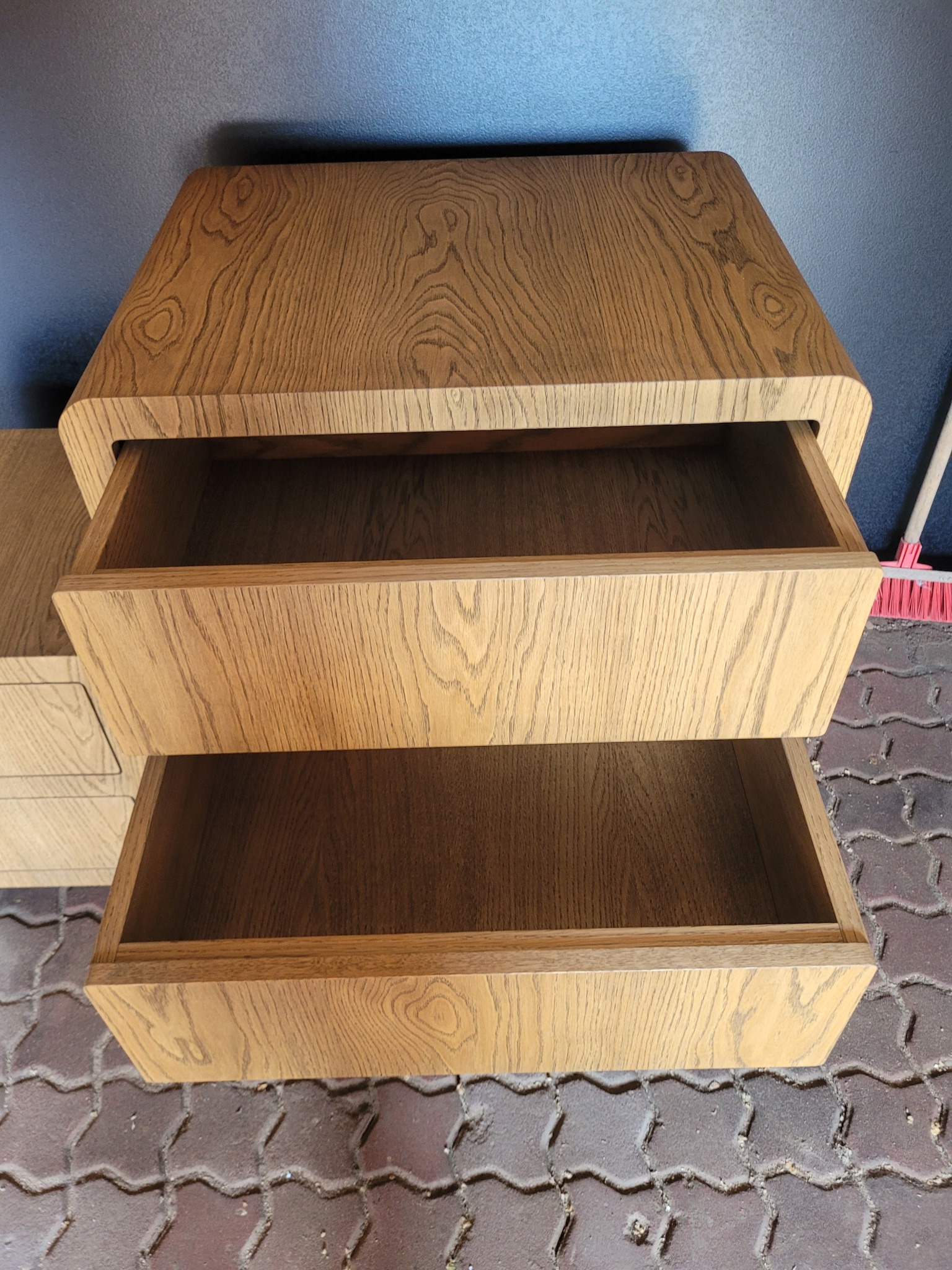 Dębowa komoda z dwoma wysuniętymi szufladami, widoczny fragment podłogi z kostki brukowej i niebieska ściana w tle.