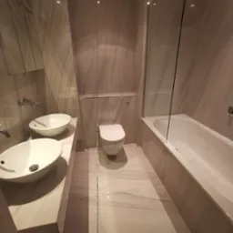 Elegancka łazienka z marmurowymi płytkami: dwie umywalki nablatowe, toaleta podwieszana i wanna za szklaną ścianką. Nowoczesny design, stonowana kolorystyka.