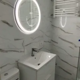 Nowoczesna łazienka z białymi płytkami imitującymi marmur, okrągłym lustrem z podświetleniem LED i grzejnikiem drabinkowym. Umywalka z szafką i toaleta w kolorze białym.