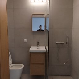 Remont expert - Nowoczesna łazienka z dużymi szarymi płytkami, podwieszaną toaletą, drewnianą szafką z umywalką i lustrem w drewnianej ramie. Szklana ścianka prysznicowa.