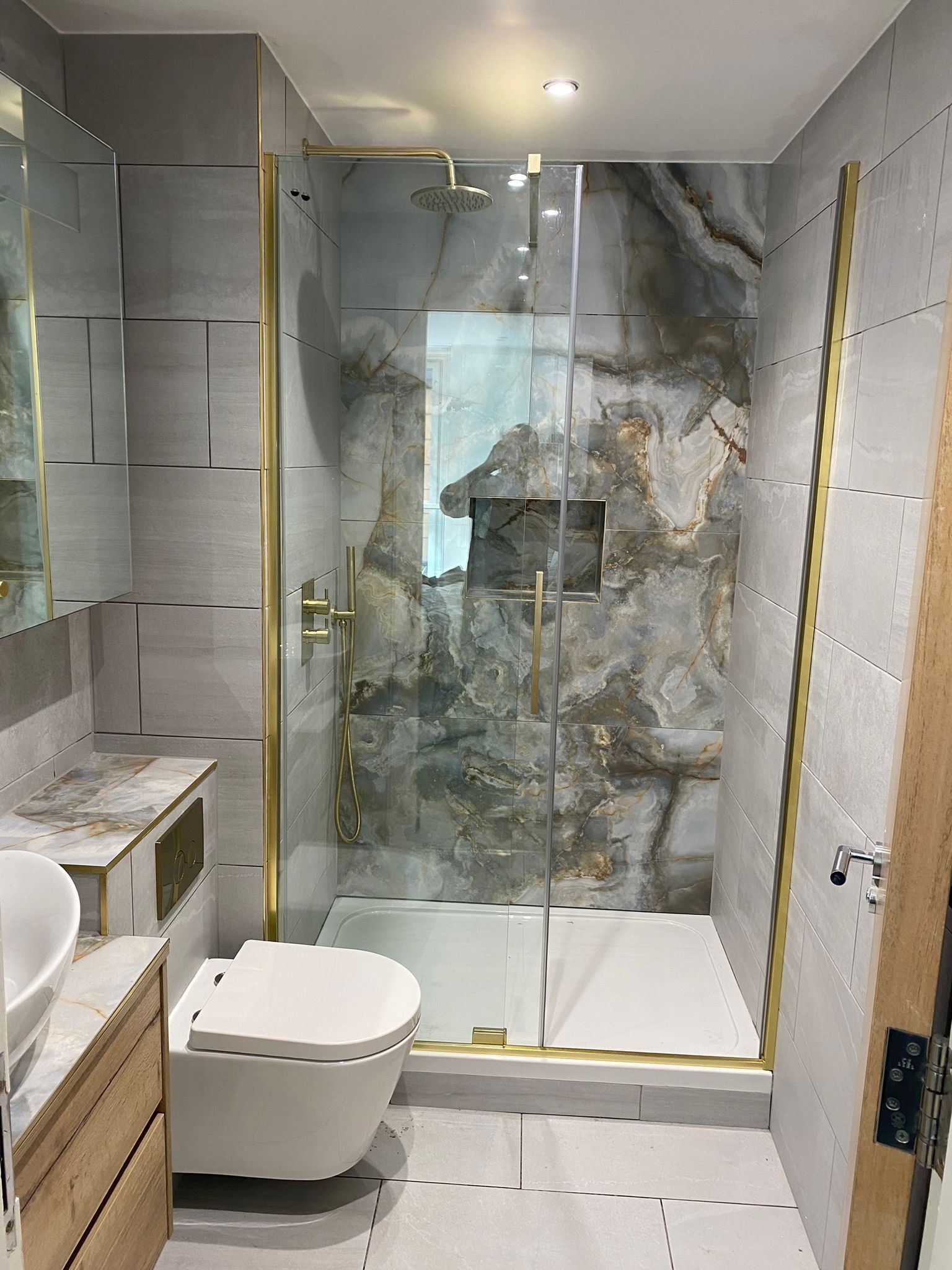 Nowoczesna łazienka z kabiną prysznicową w złotej ramie, płytki imitujące kamień, wisząca toaleta i umywalka nablatowa.