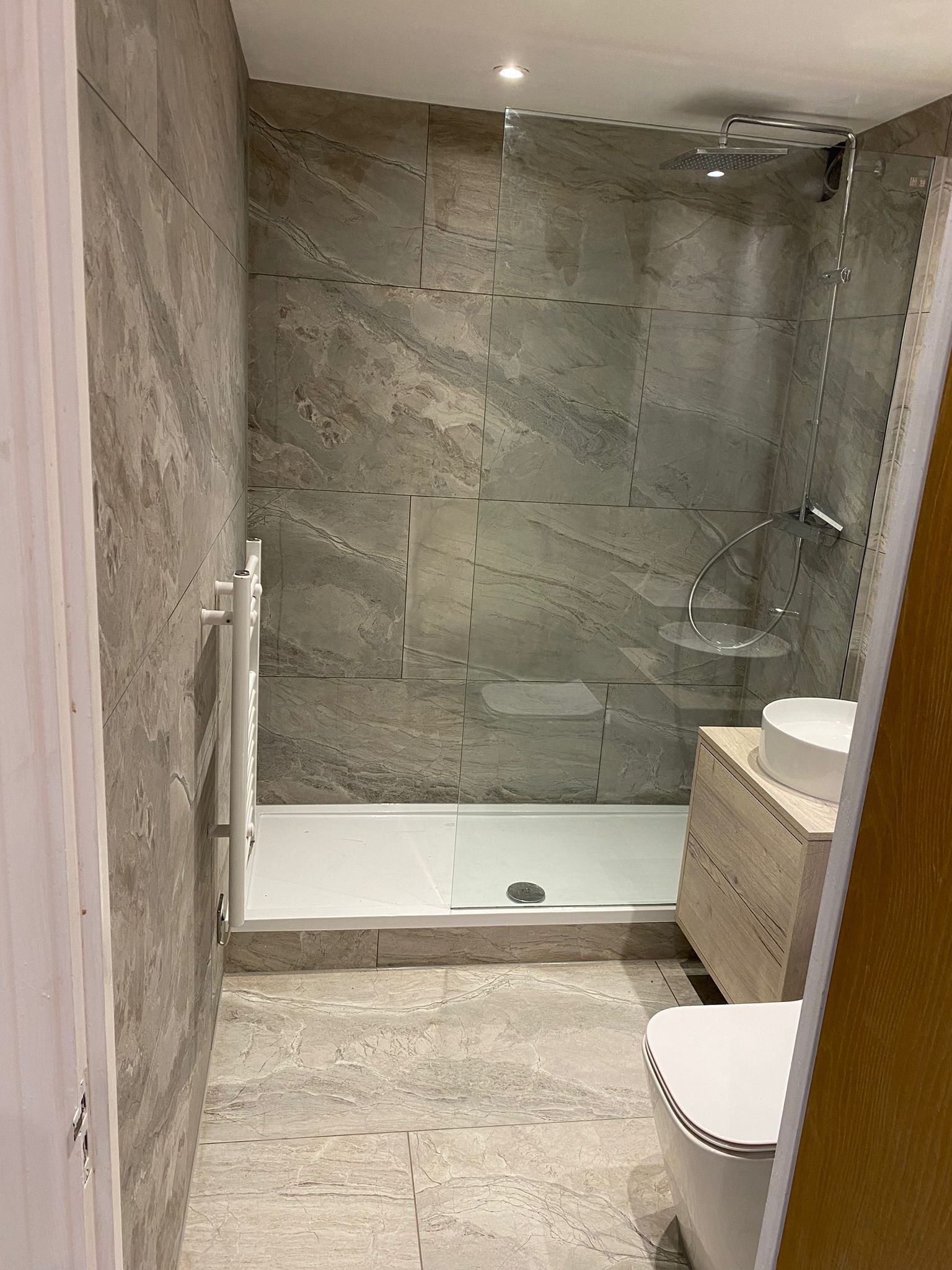 Nowoczesna łazienka z kabiną prysznicową typu walk-in, umywalką nablatową na drewnianej szafce i grzejnikiem drabinkowym. Szare płytki imitujące marmur na ścianach i podłodze.