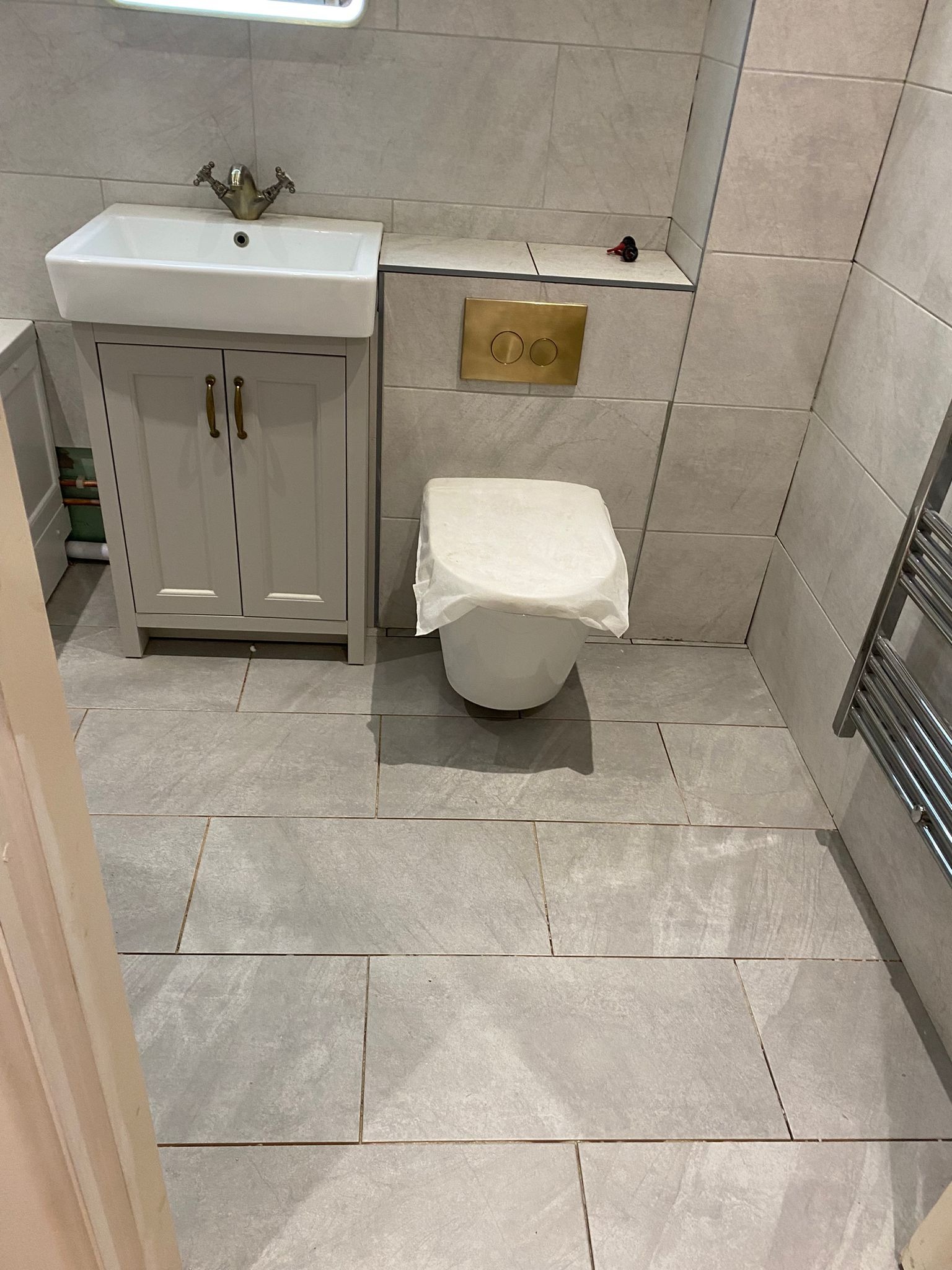 Wykończona łazienka z szarymi płytkami, umywalką z szafką w stylu retro i złotymi akcentami. Toaleta zabezpieczona folią, grzejnik drabinkowy po prawej stronie.