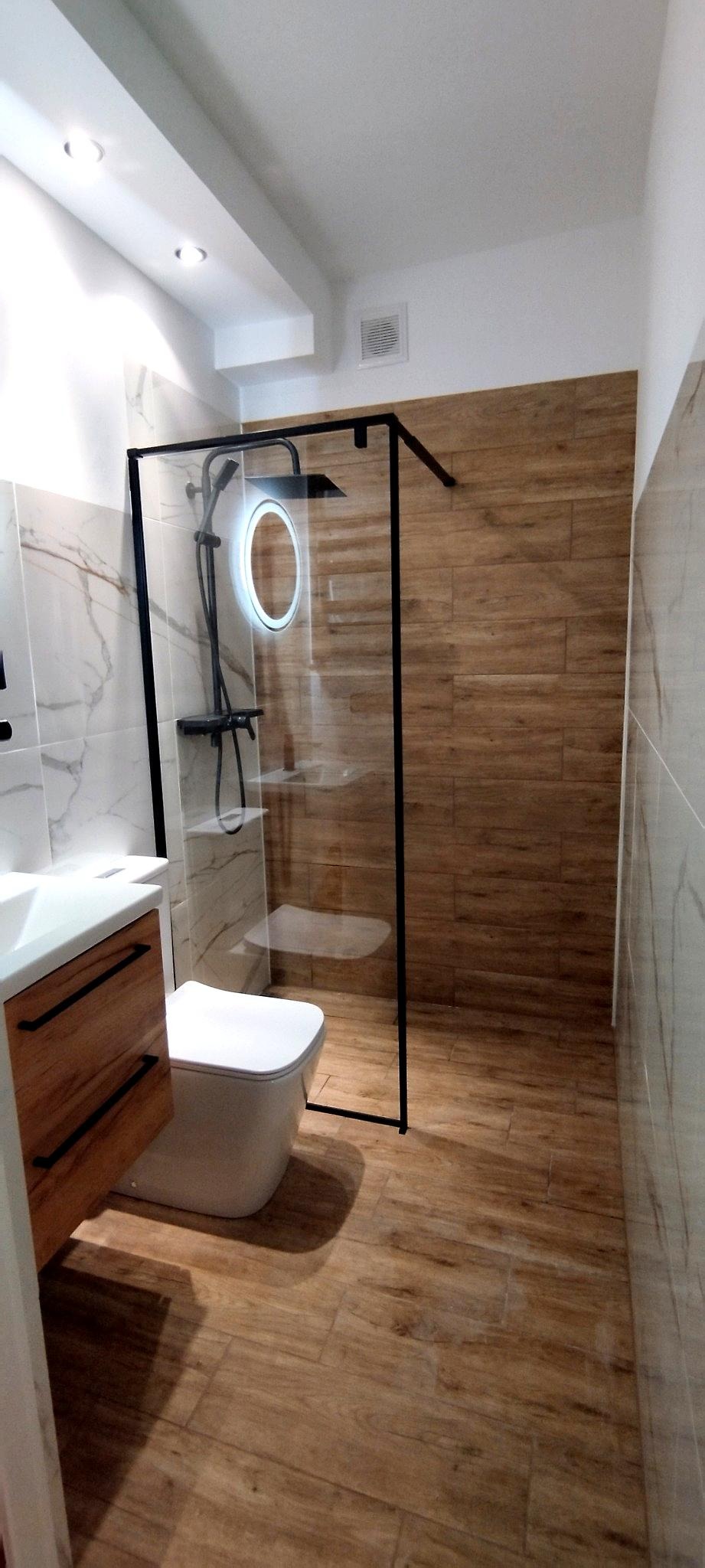 Nowoczesna łazienka z kabiną prysznicową typu walk-in, wykończona płytkami imitującymi drewno i marmur, z czarną armaturą i oświetleniem LED.