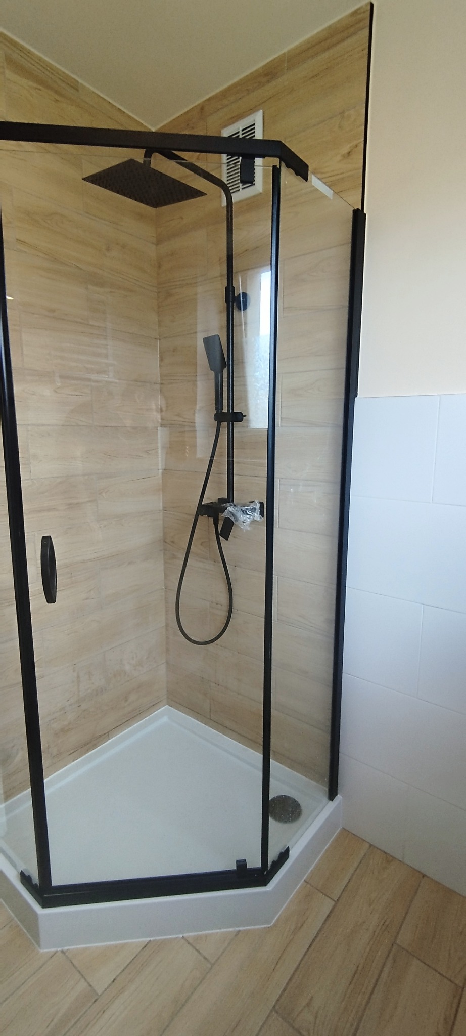 Wykończona kabina prysznicowa z czarnymi ramami i deszczownicą, obłożona płytkami imitującymi drewno, z widocznym odpływem liniowym. Nowoczesna aranżacja łazienki.