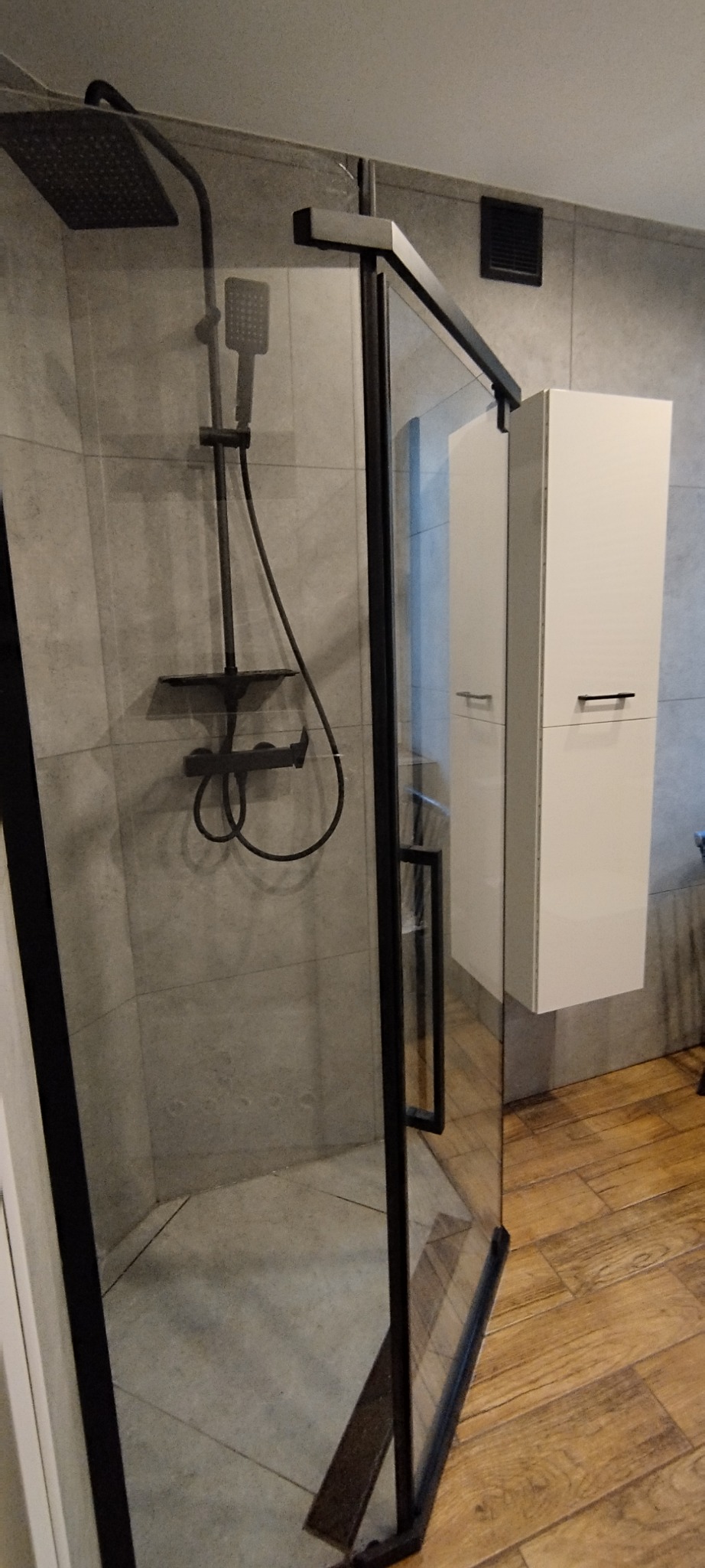 Nowoczesna kabina prysznicowa z czarnymi profilami i deszczownicą, szare płytki na ścianach, drewnopodobna podłoga, minimalistyczna szafka łazienkowa.