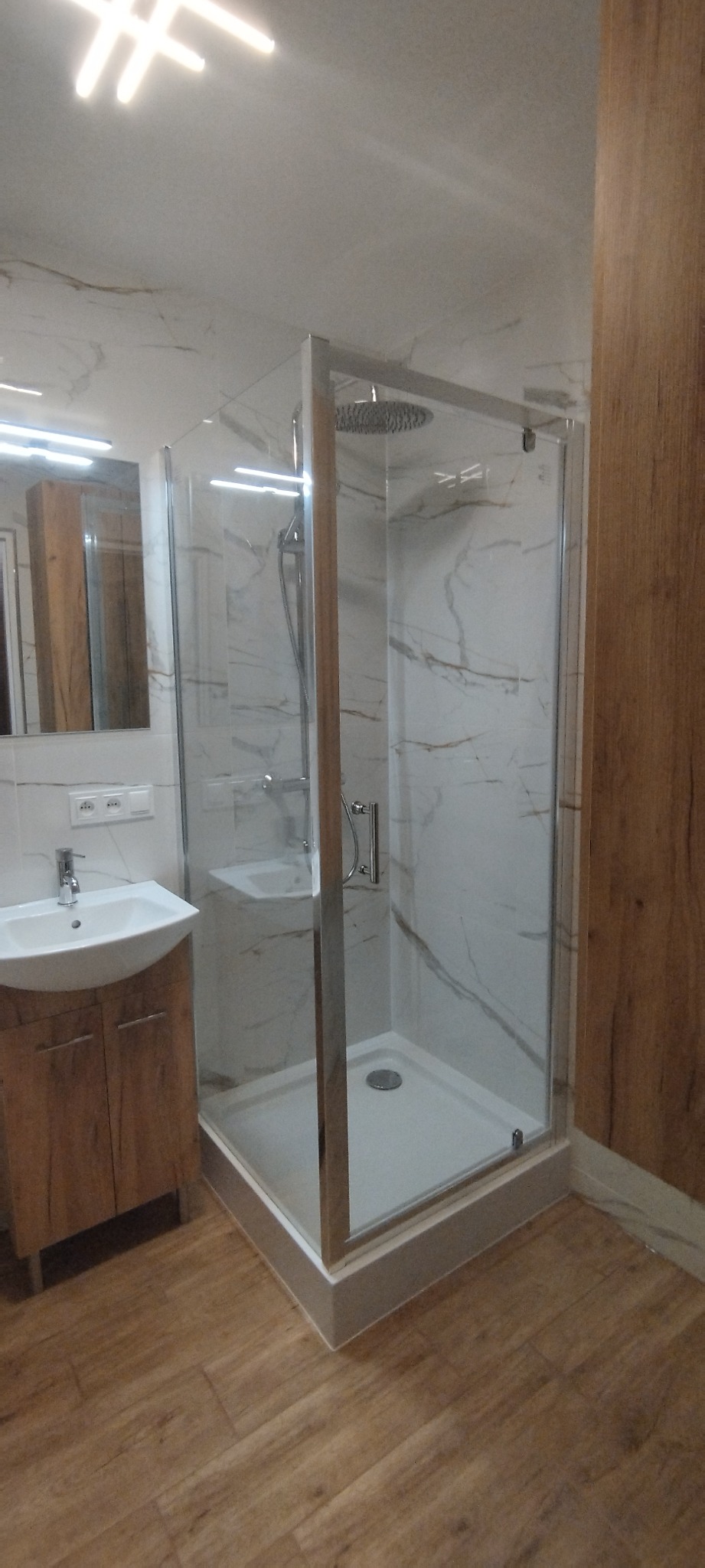 Nowoczesna kabina prysznicowa z deszczownicą, umywalka z szafką w wykończonej łazience. Płytki imitujące marmur na ścianach i drewnopodobne na podłodze.