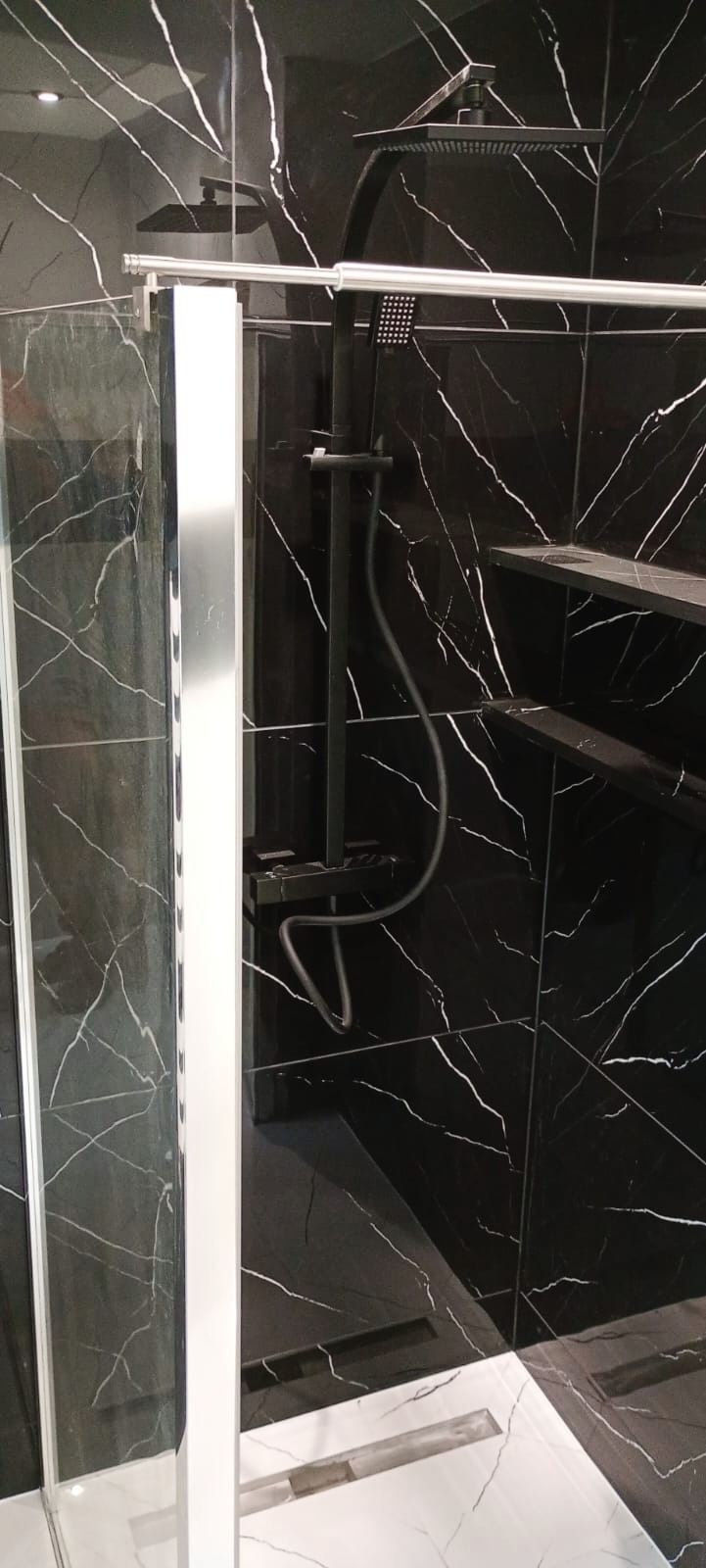 Nowoczesna kabina prysznicowa z czarnymi płytkami imitującymi marmur z białymi żyłami, czarna armatura i biała podłoga z odpływem liniowym.